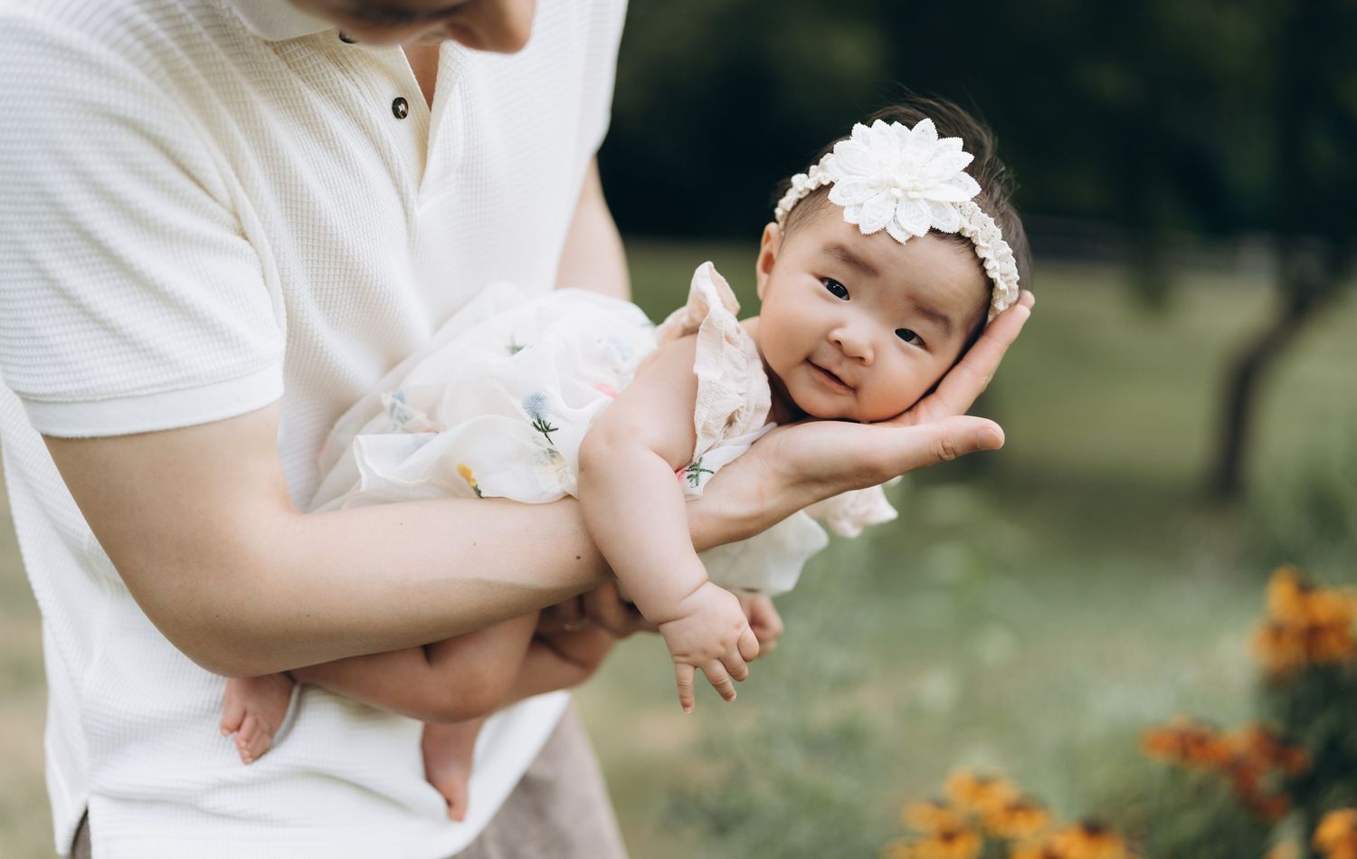Adorable baby wearing white dress and floral headband smiles while being held outdoors near orange flowers.