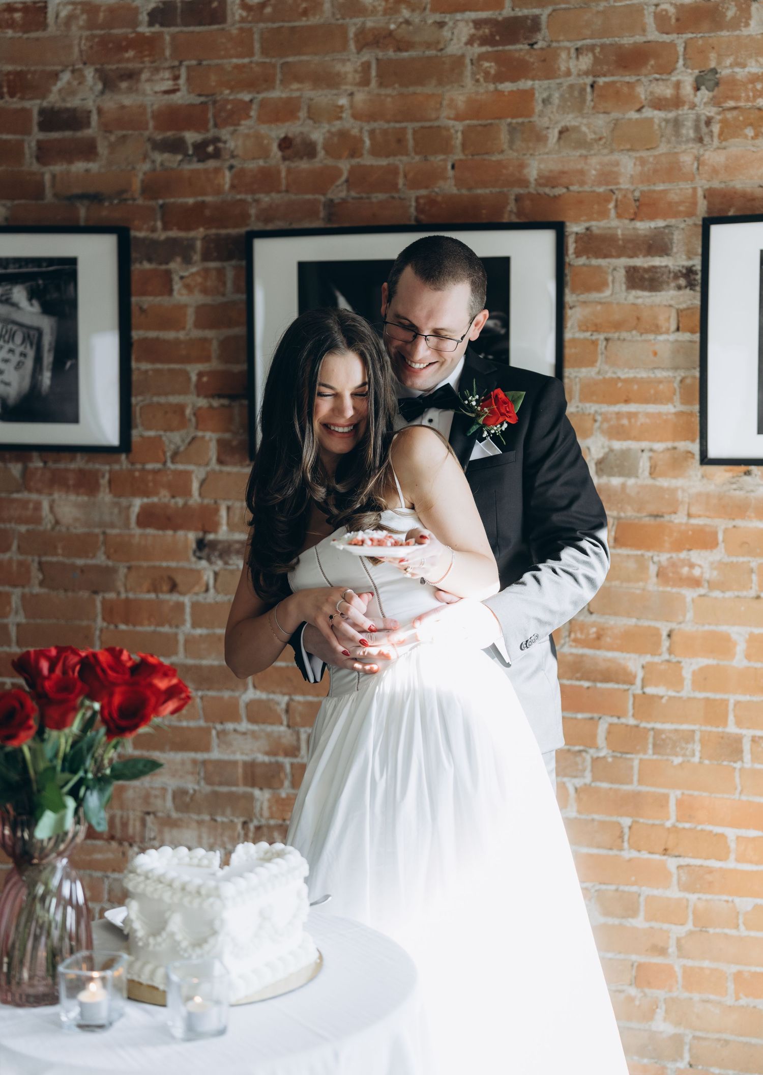 Intimate newlywed moment during wedding reception against exposed brick wall with white and red floral accents.