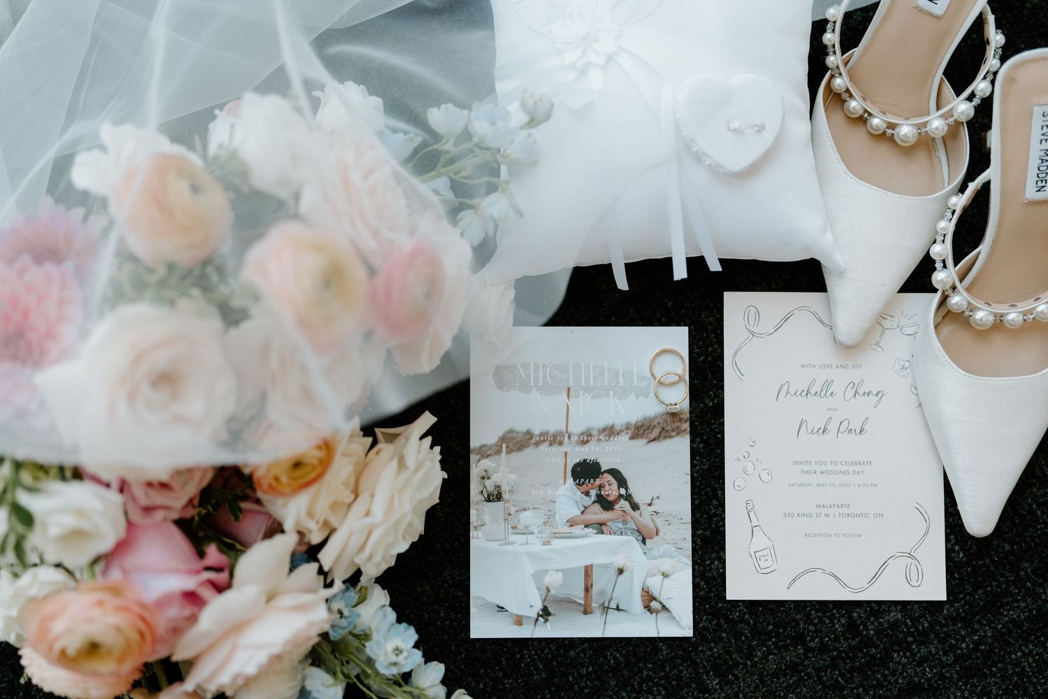 Wedding details flatlay with invitation cards, white heels, and soft pink roses on grey background.