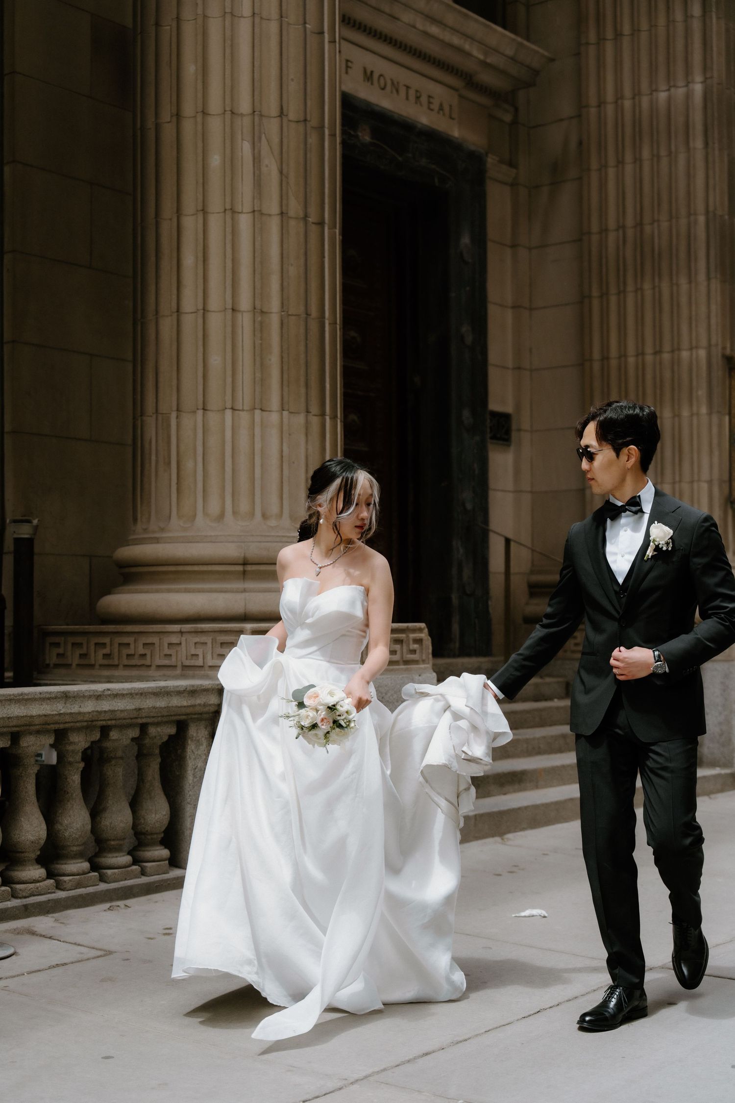 Elegant couple in formal wedding attire walking together outside a grand stone building in Montreal.