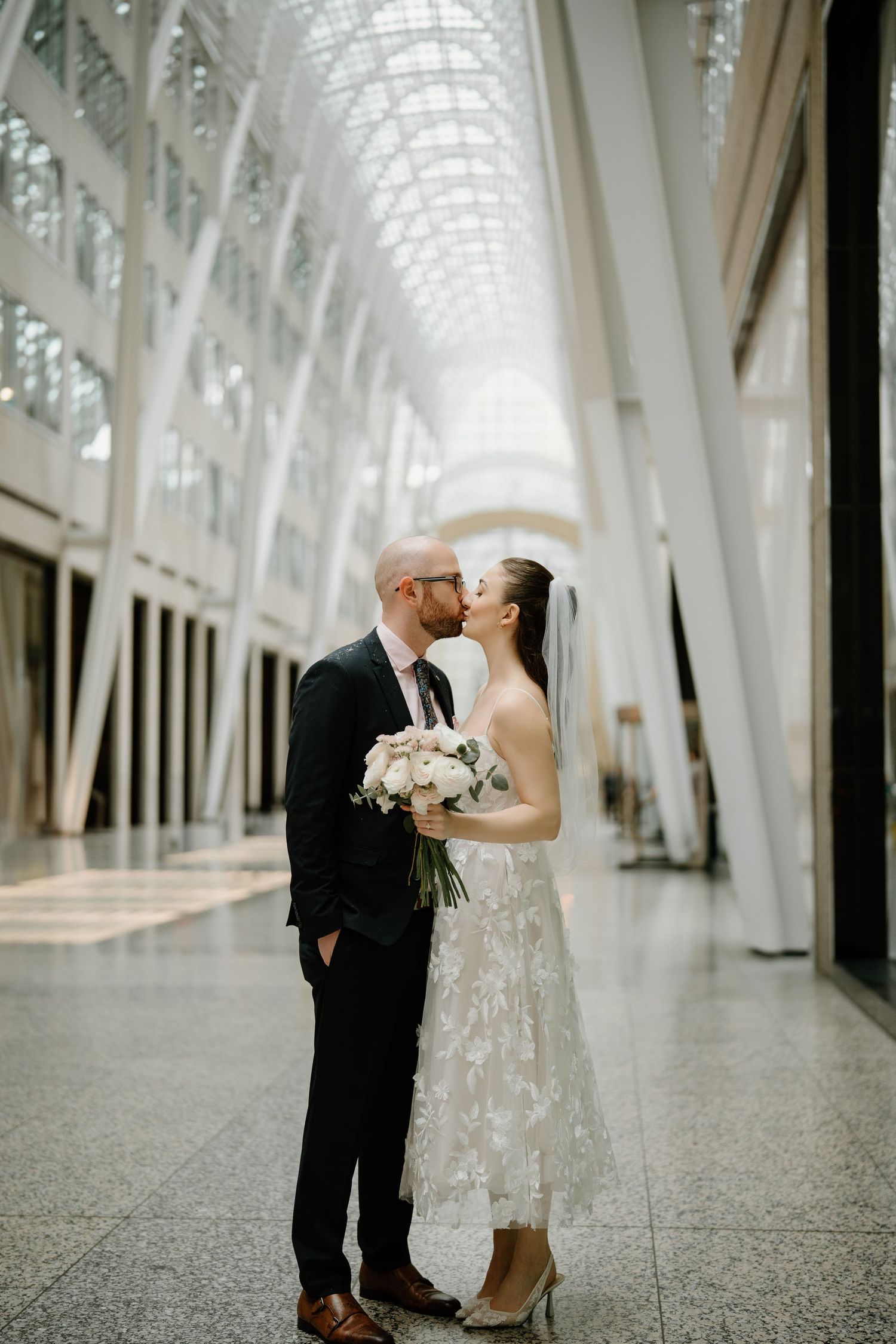 Couple embraces in an elegant architectural space with soaring glass ceiling and white columns creating a dramatic backdrop.