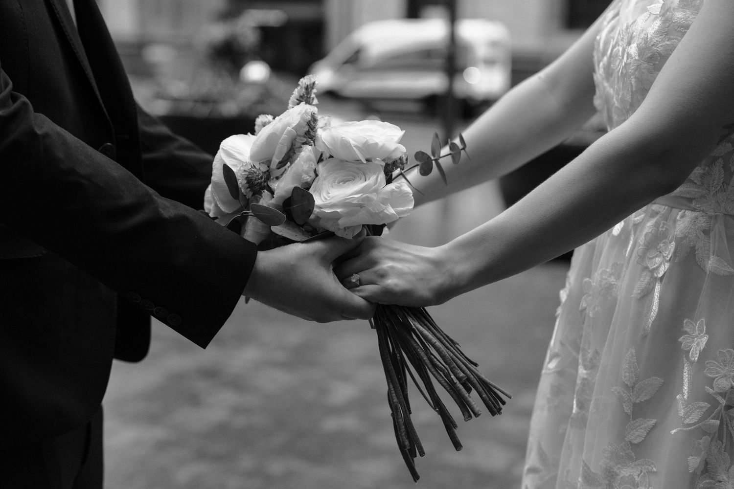 A black and white close-up of hands exchanging a wedding bouquet during a wedding ceremony.