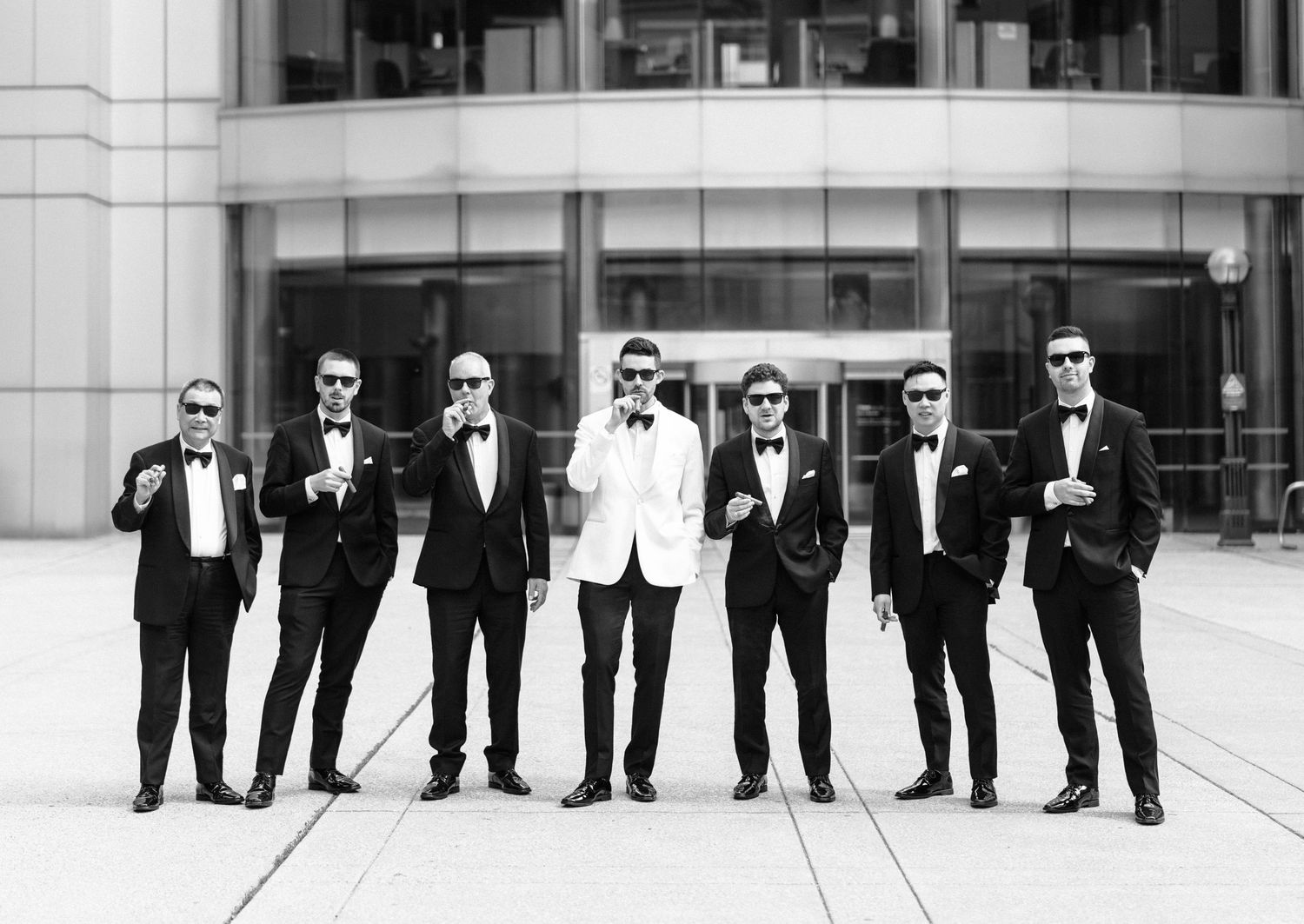 A group of seven men in formal attire with sunglasses standing in a line outside a modern building.