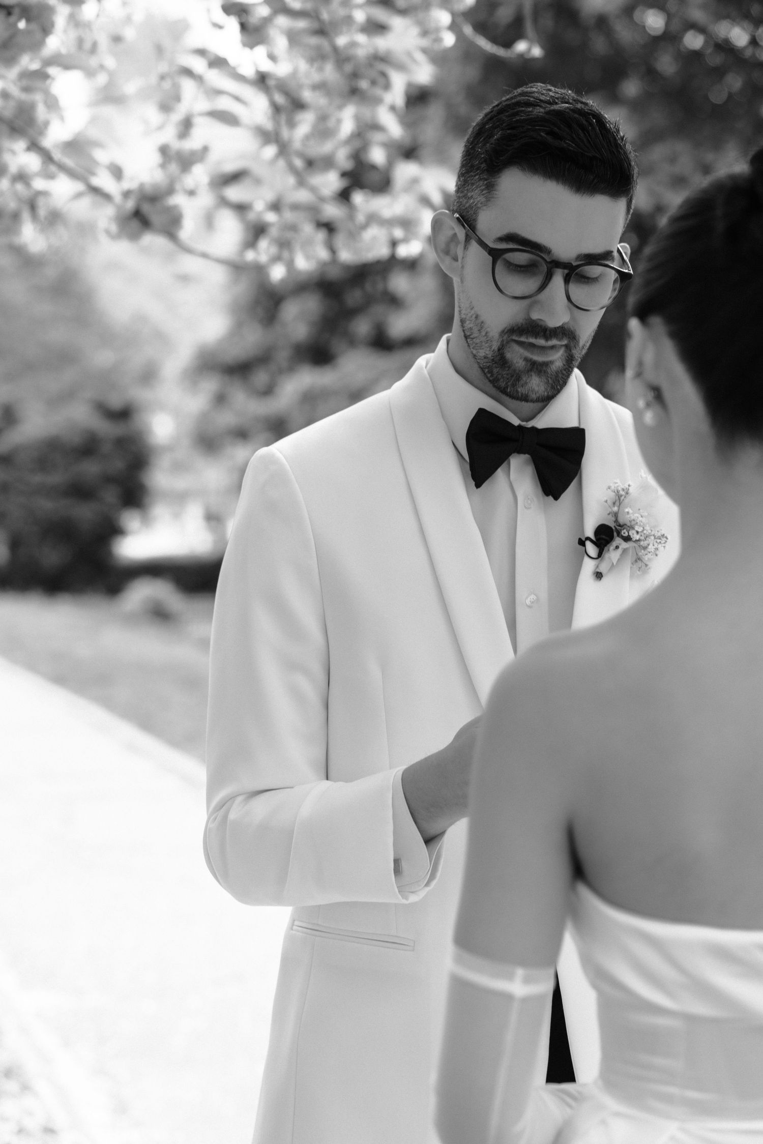 A black and white wedding photograph showing a groom in a white tuxedo jacket and bowtie during an intimate ceremony moment.