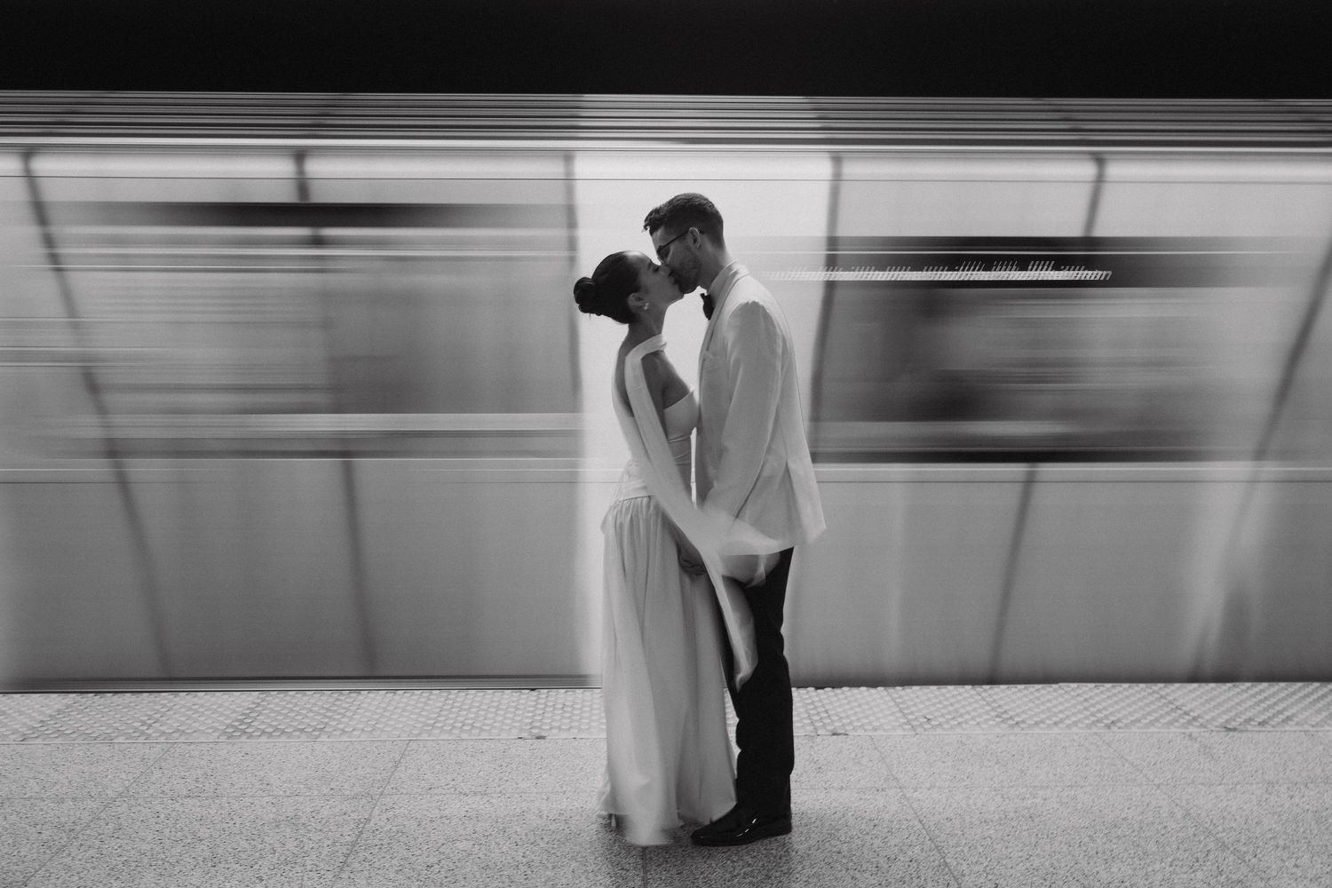 A couple in formal wear share an intimate moment in a subway station while a train blurs past in black and white.