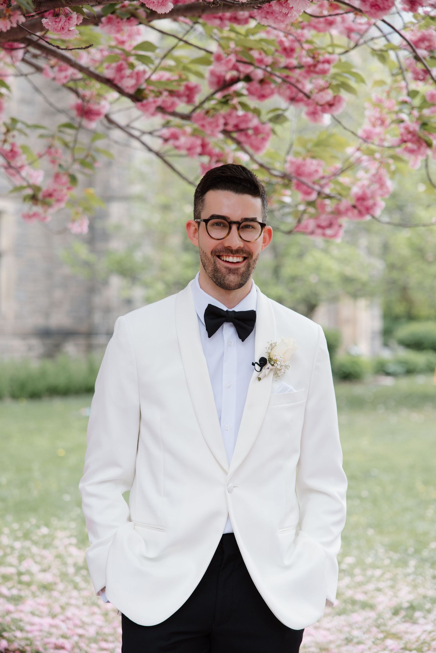A groom in a white dinner jacket and black bow tie stands under blooming pink cherry blossoms in a garden setting.