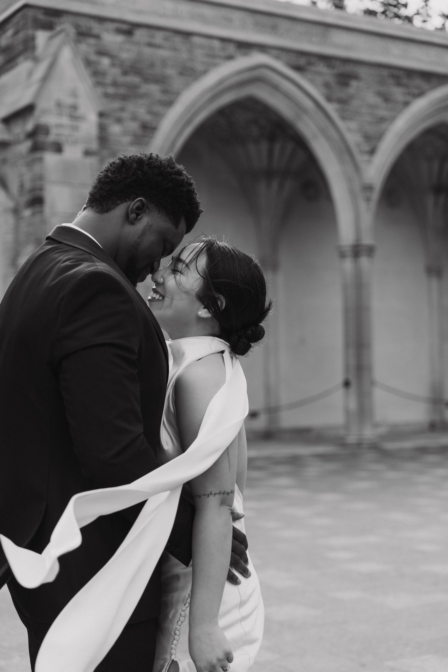 A romantic black and white wedding photo in front of stone Gothic arches showing a couple in an intimate embrace.