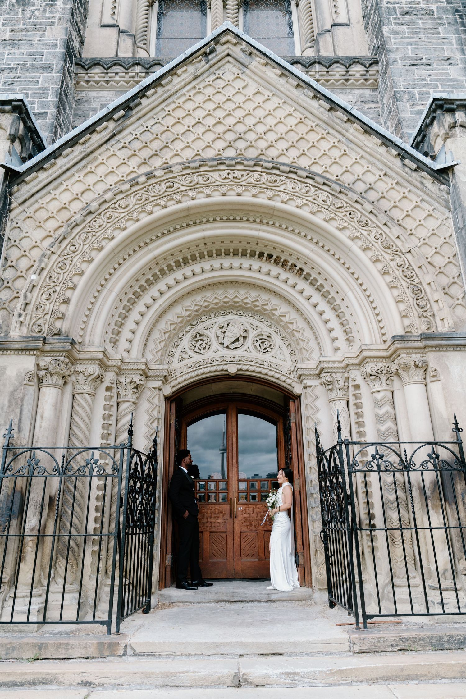 Elegant stone church entrance with ornate archway and decorative ironwork gates showing two figures in formal attire.