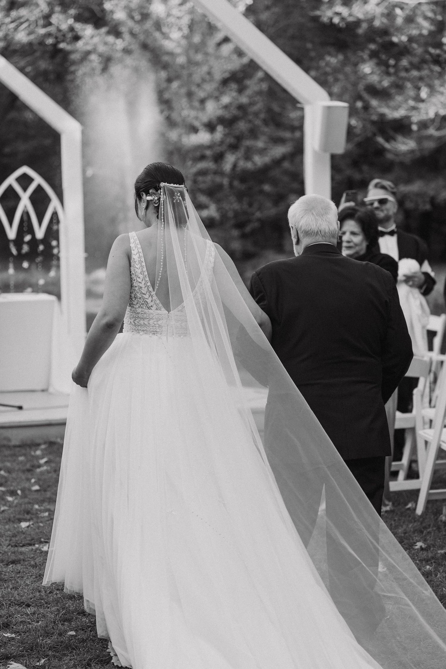 Back view of bride in lace wedding dress with flowing veil walking towards outdoor chapel.