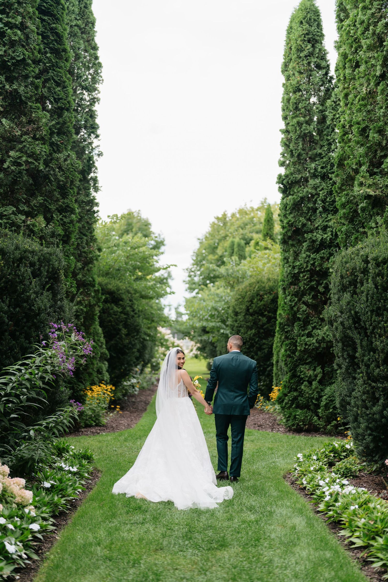 A couple walks down a scenic garden path lined with tall cypress trees and flowering bushes.