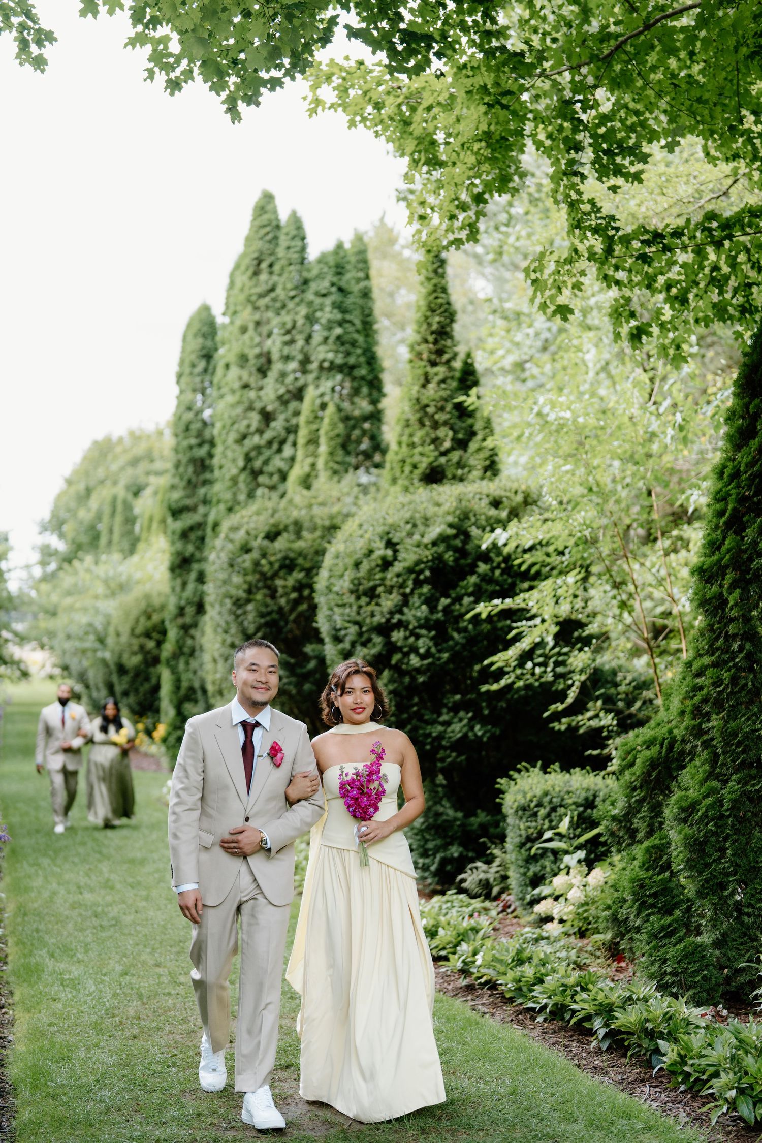 A couple walks together in a lush garden setting with tall evergreen trees and greenery along a path.