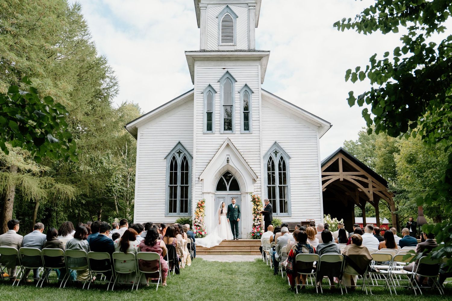 A charming white country church with a tall steeple stands surrounded by trees during an outdoor wedding ceremony.