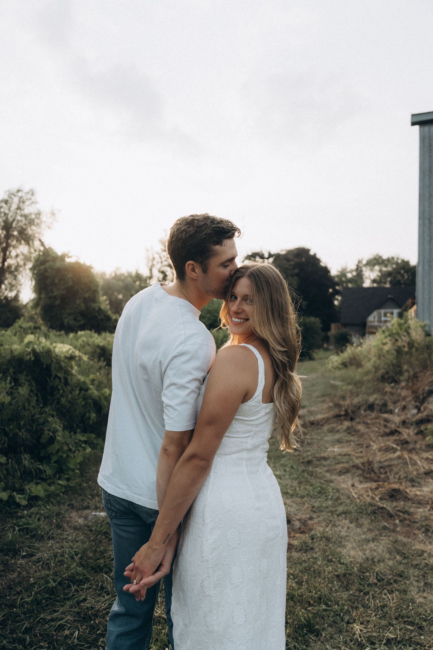 Couple in white and casual attire share an intimate moment on a grassy hillside at sunset.