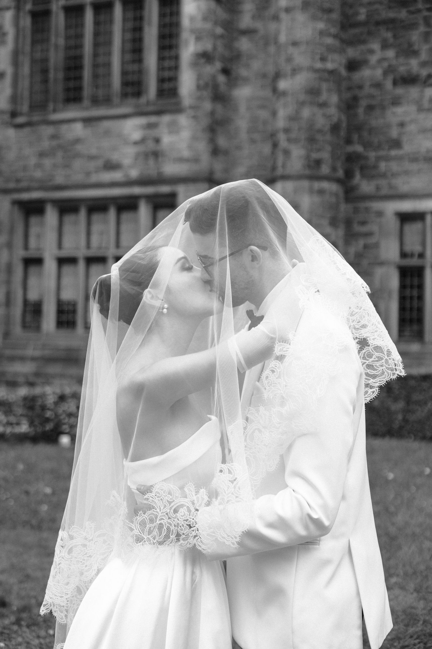 A romantic black and white wedding photo showing a tender moment under a flowing veil at a historic stone building.