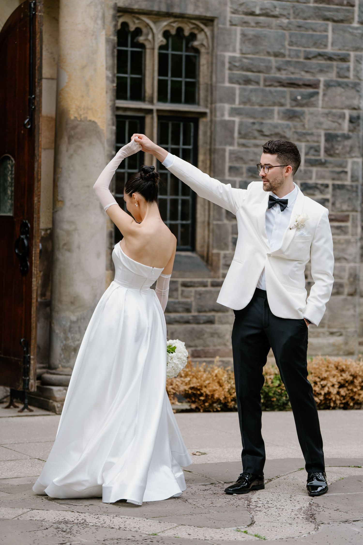 A bride in a white wedding gown and groom in white tuxedo strike playful dance poses against a stone church exterior.