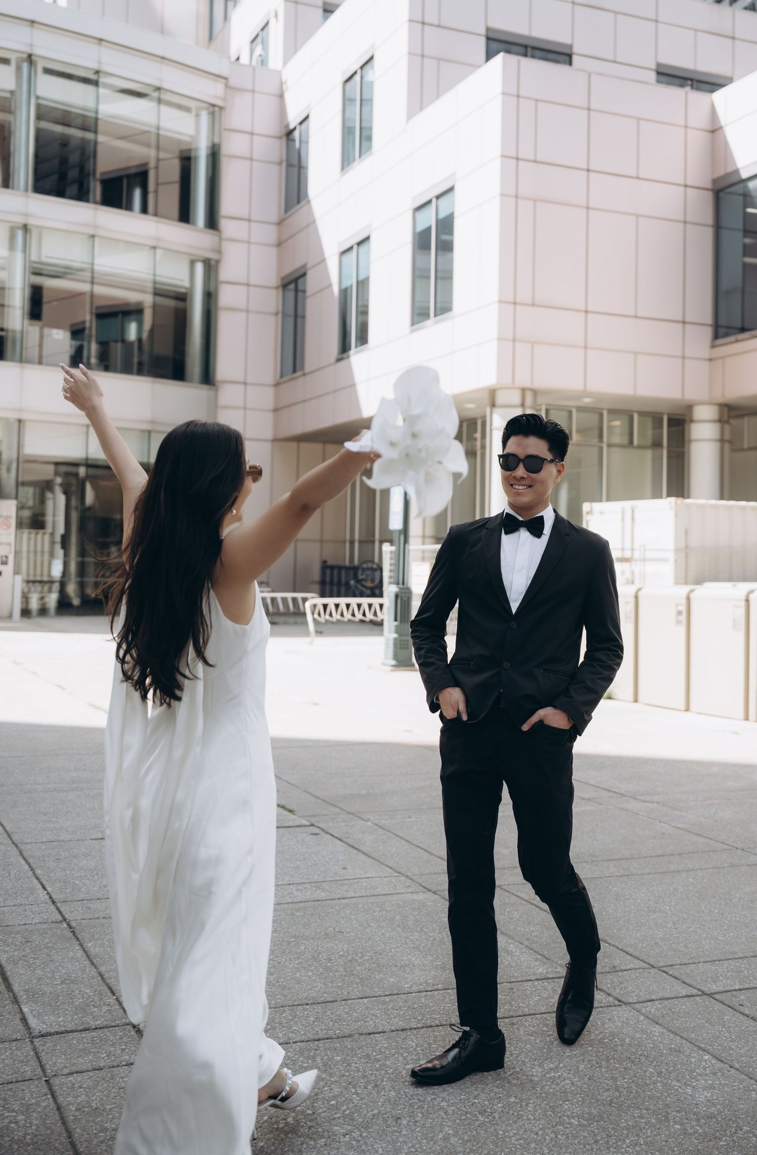 A couple in wedding attire poses in front of a modern building with white exterior and glass windows.