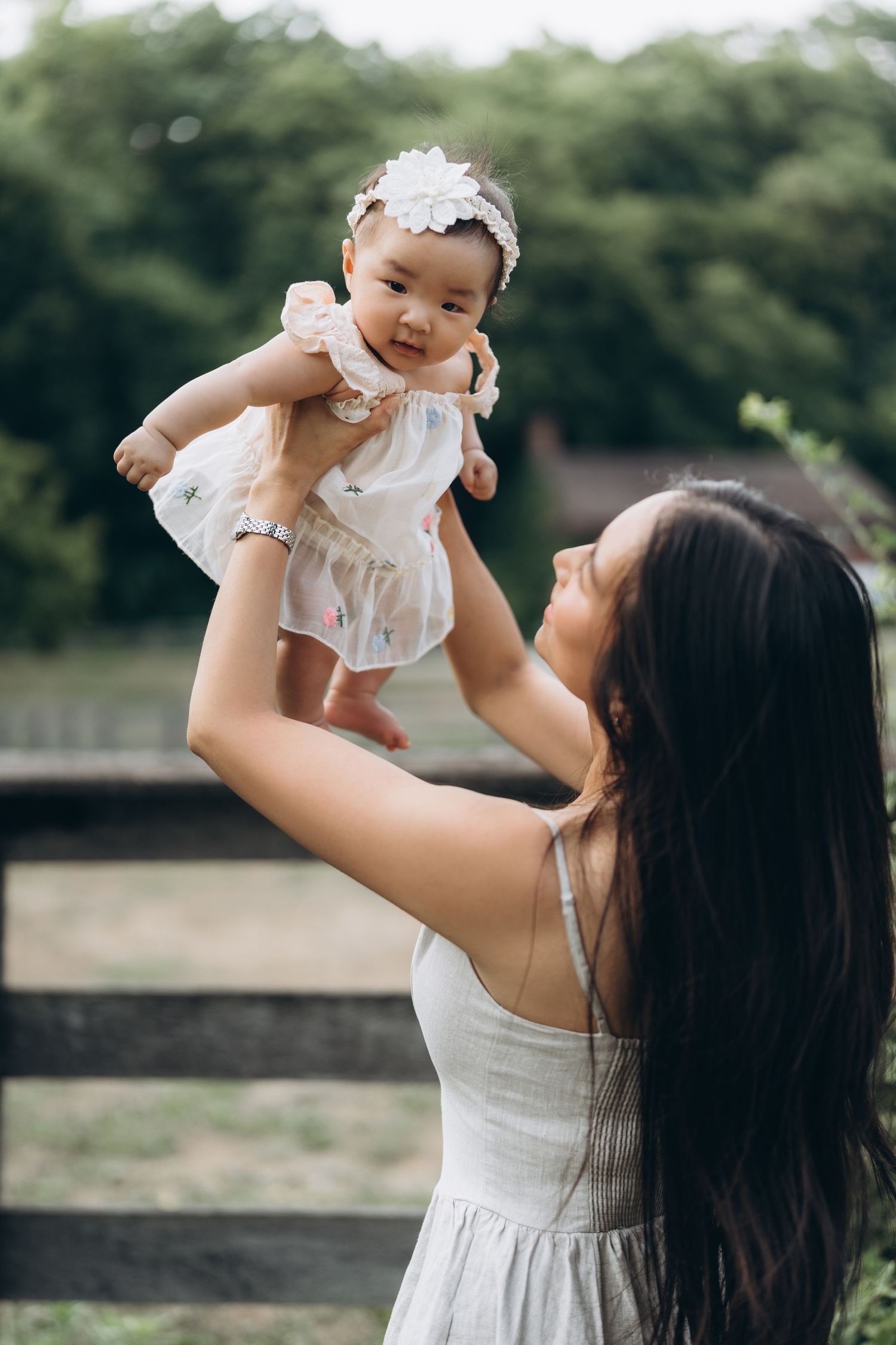 A tender outdoor moment of affection between parent and baby in white outfits against a natural background.