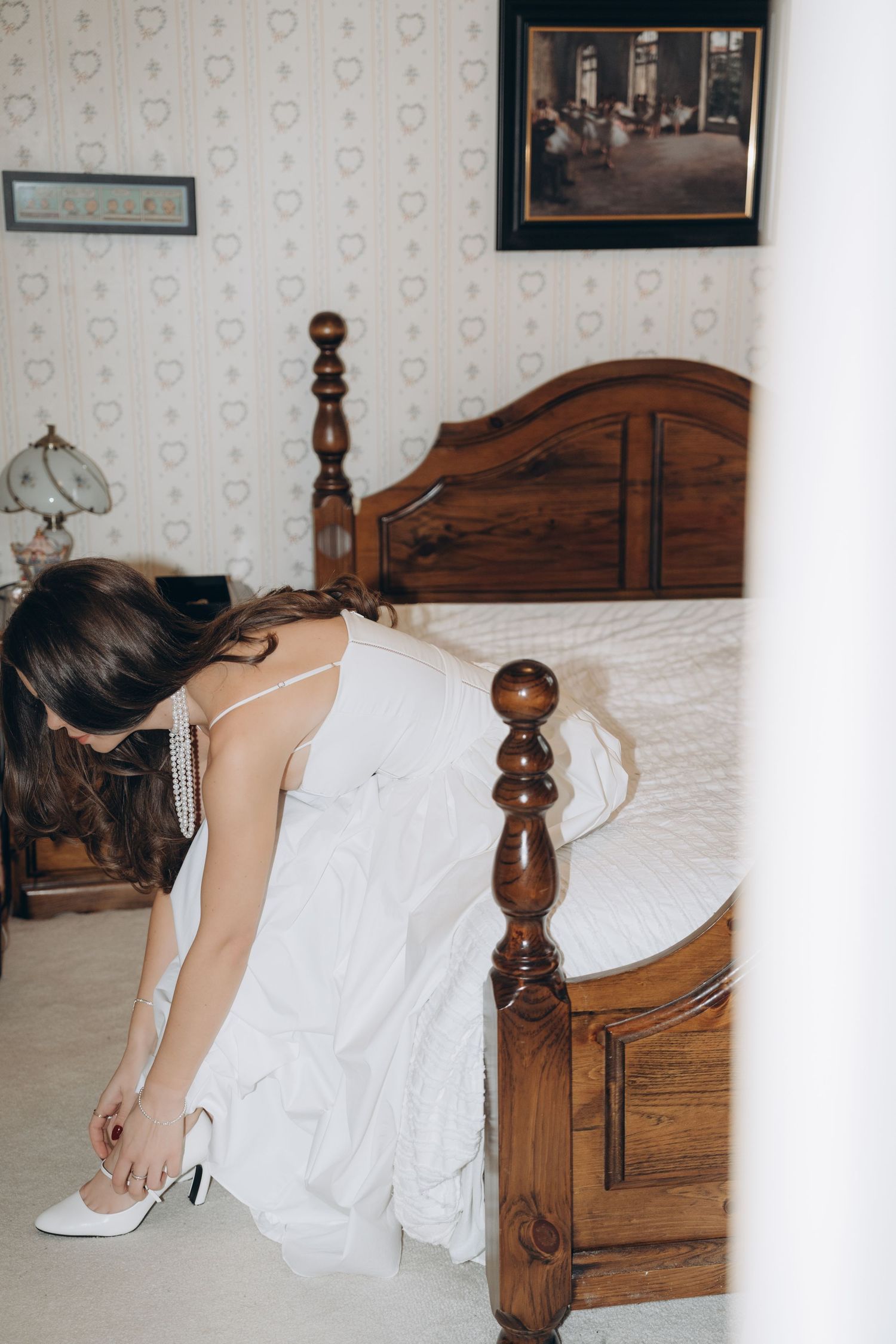 Person in a white wedding gown sitting on an antique wooden bed, showing detail of the dress and shoes.