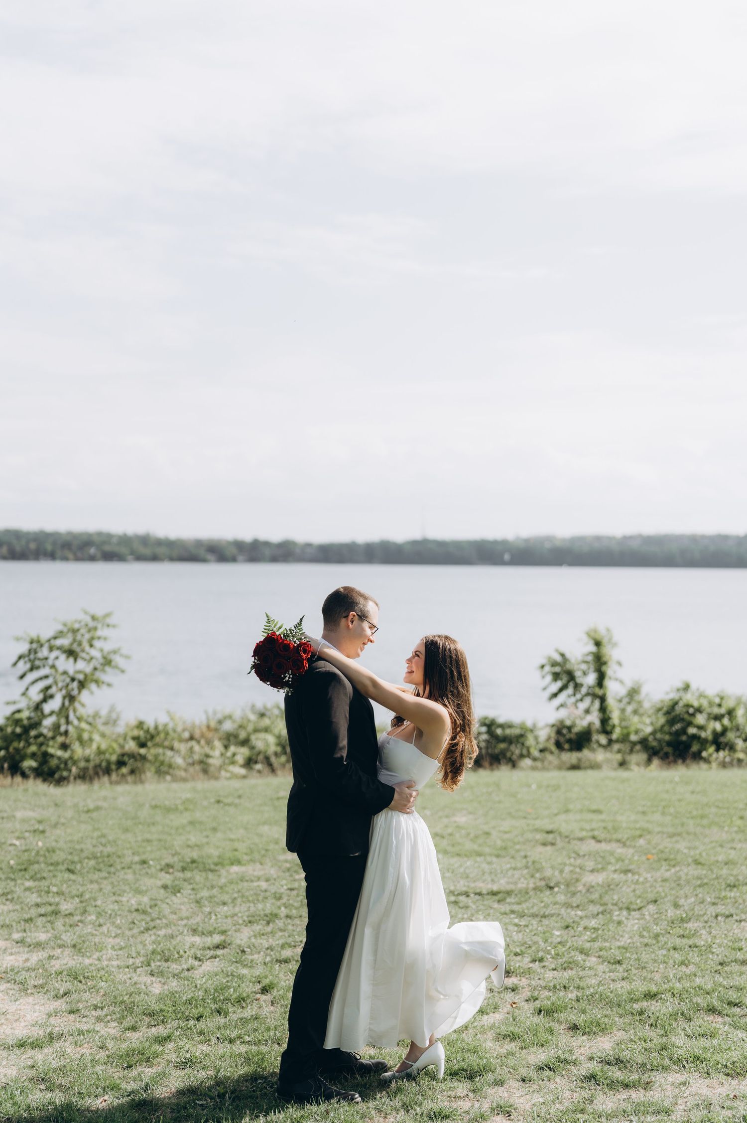 A couple in wedding attire dance on a grassy overlook with a serene lake view in the background.