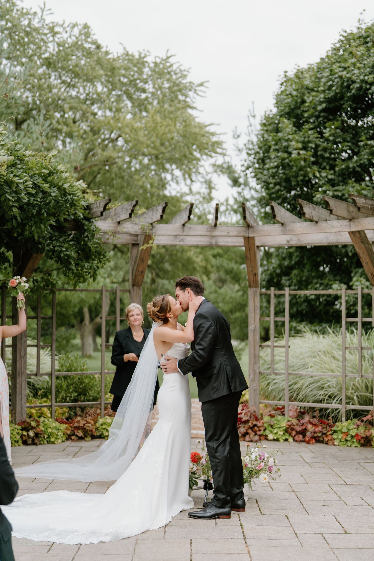 A romantic wedding ceremony kiss under a rustic wooden pergola surrounded by lush garden greenery.