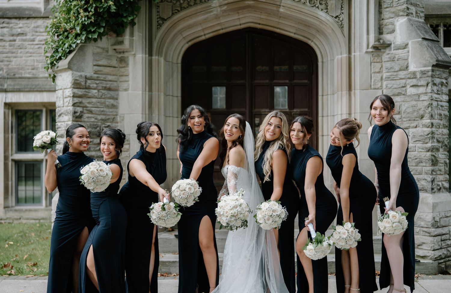 Wedding party in black dresses poses with bride in white gown outside stone church building.