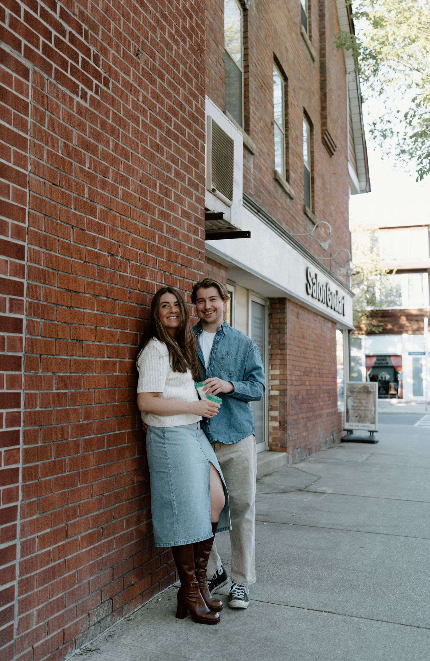 A couple stands together outside a brick building with storefronts on a sunny day.