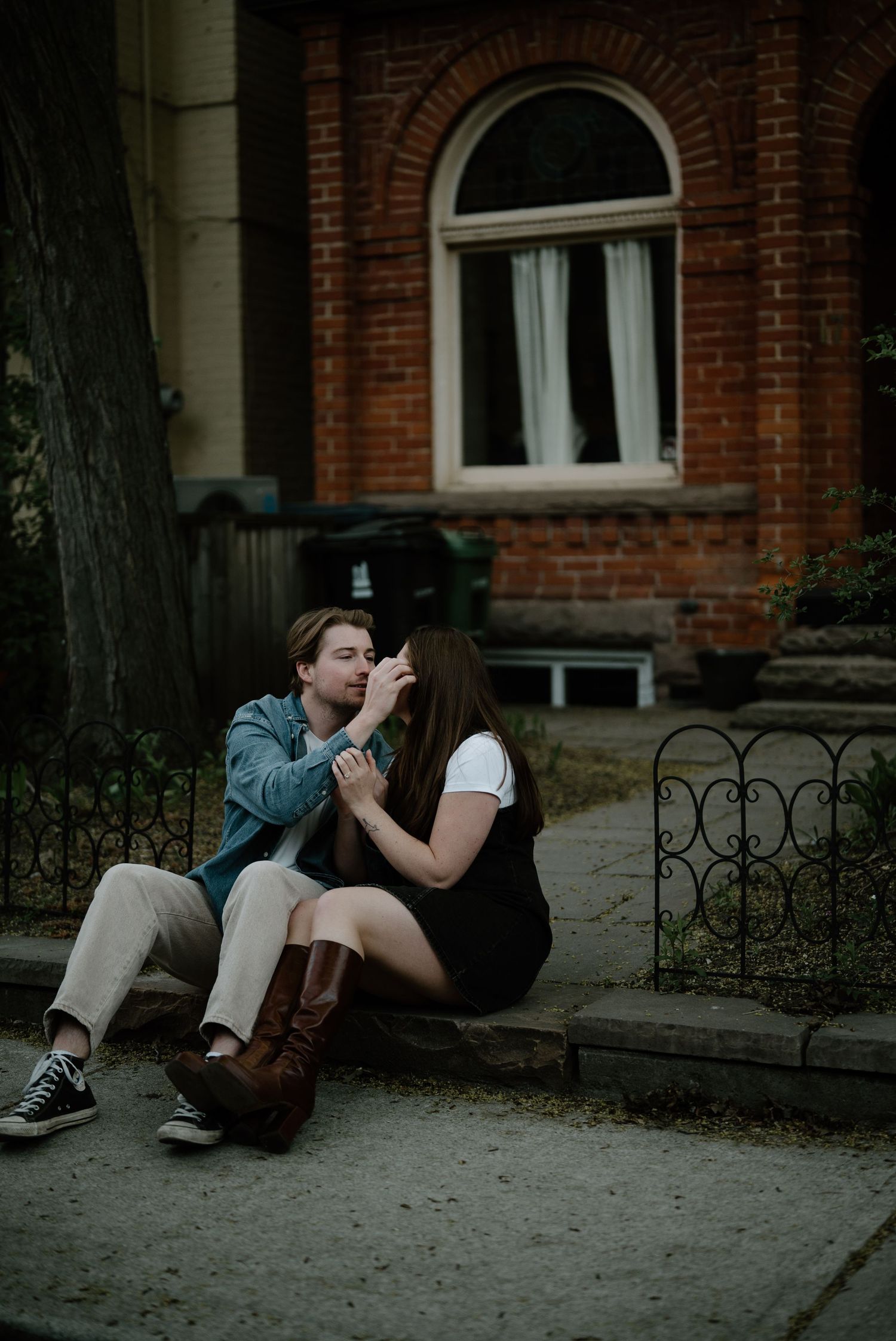 A romantic couple sits together on stairs outside a brick building with an arched window and iron fence at dusk.