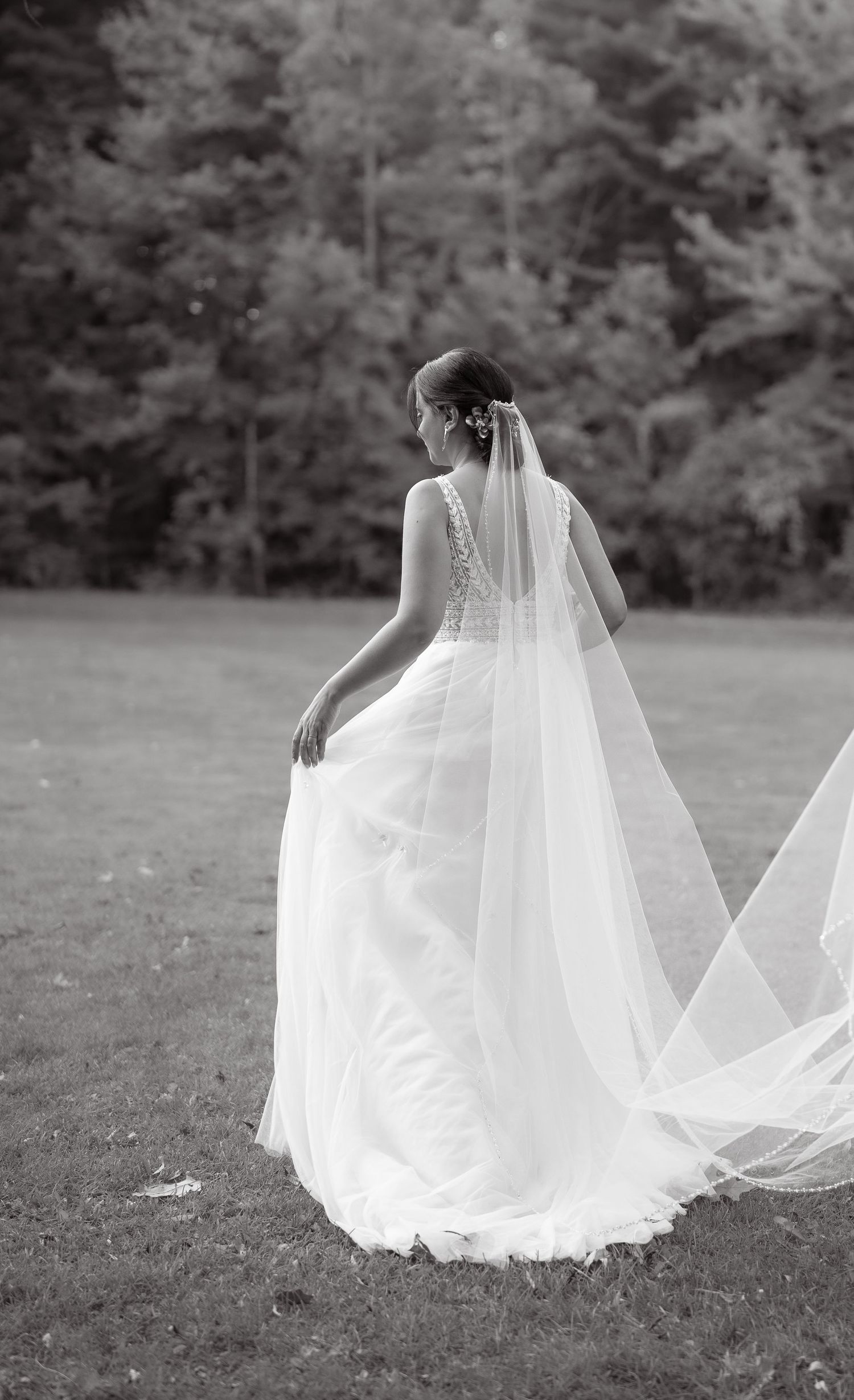 Black and white portrait of bride in lace dress with flowing veil against forest backdrop.