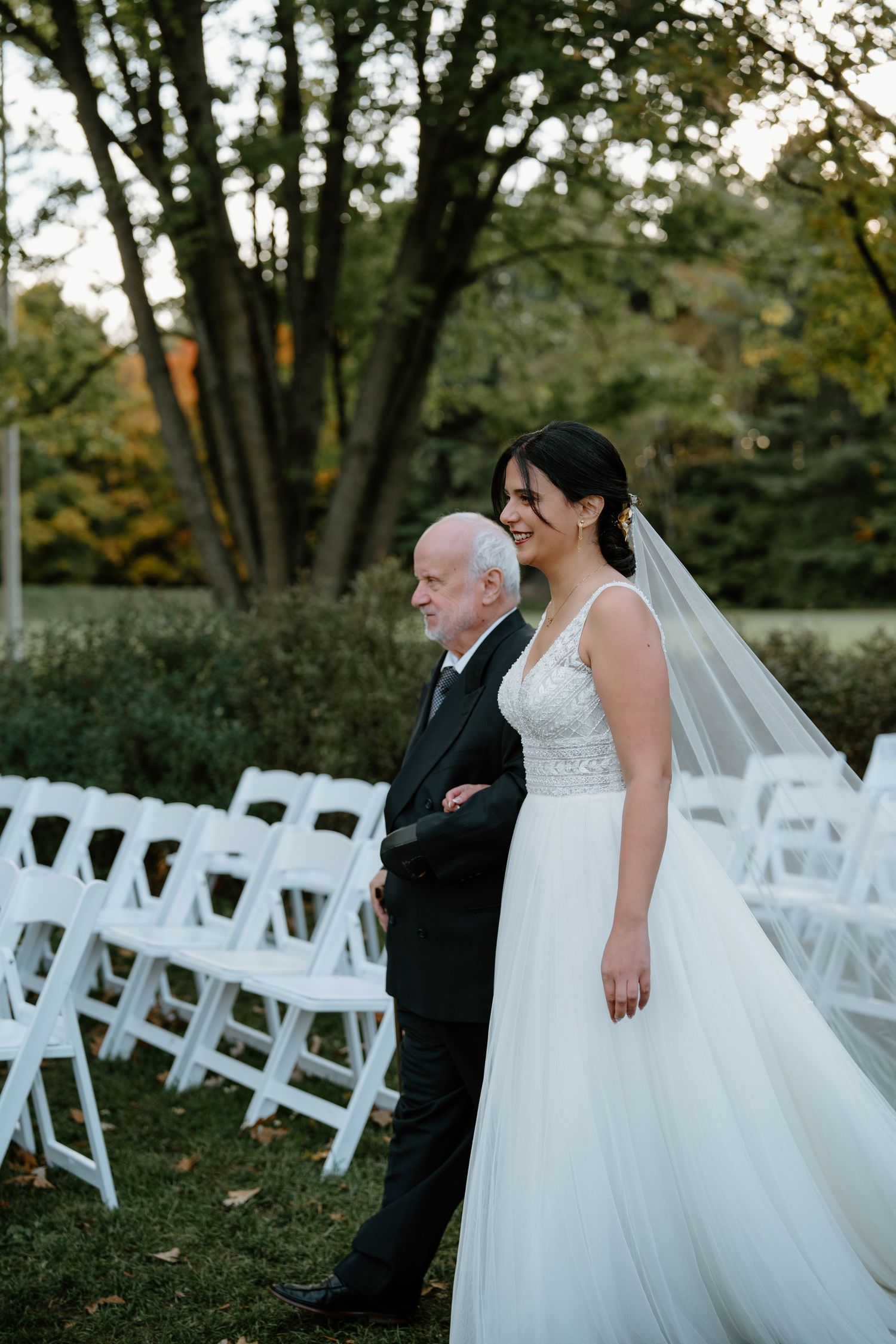 A touching processional moment before white guest chairs at an outdoor autumn wedding.