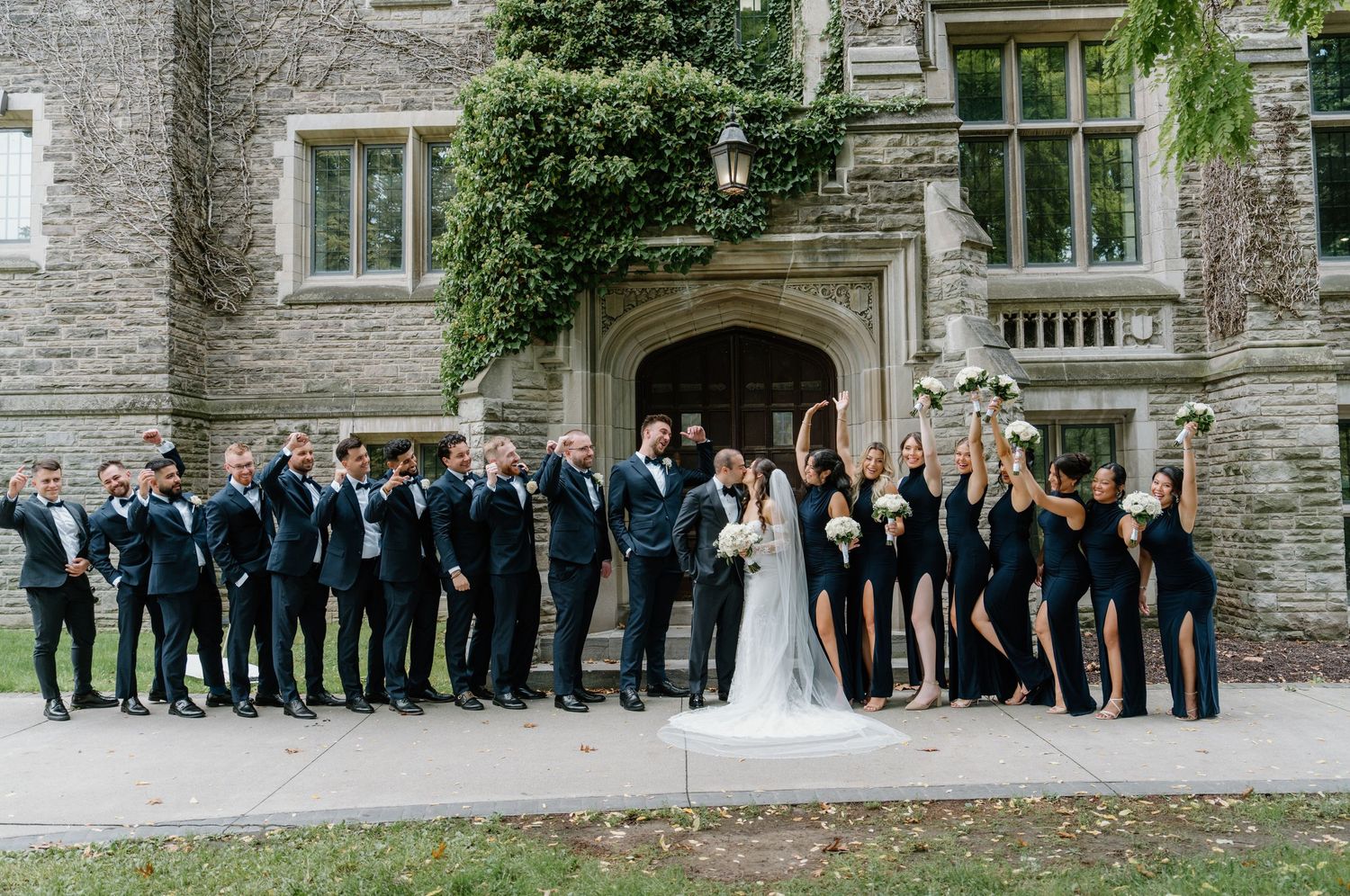 A wedding party poses in front of a stone building exterior in formal attire.