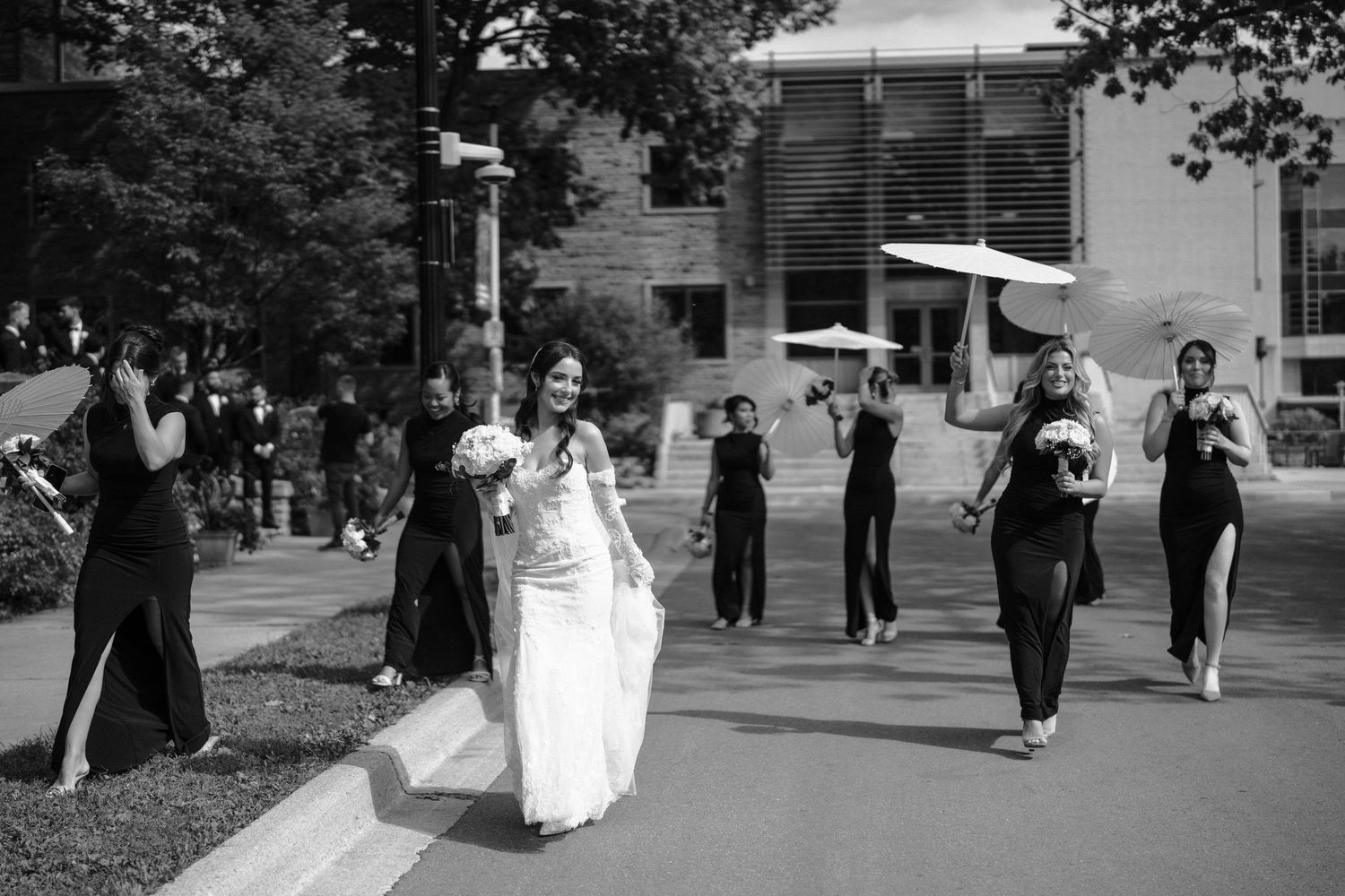 Wedding party walks together holding white umbrellas and bouquets along a tree-lined path.