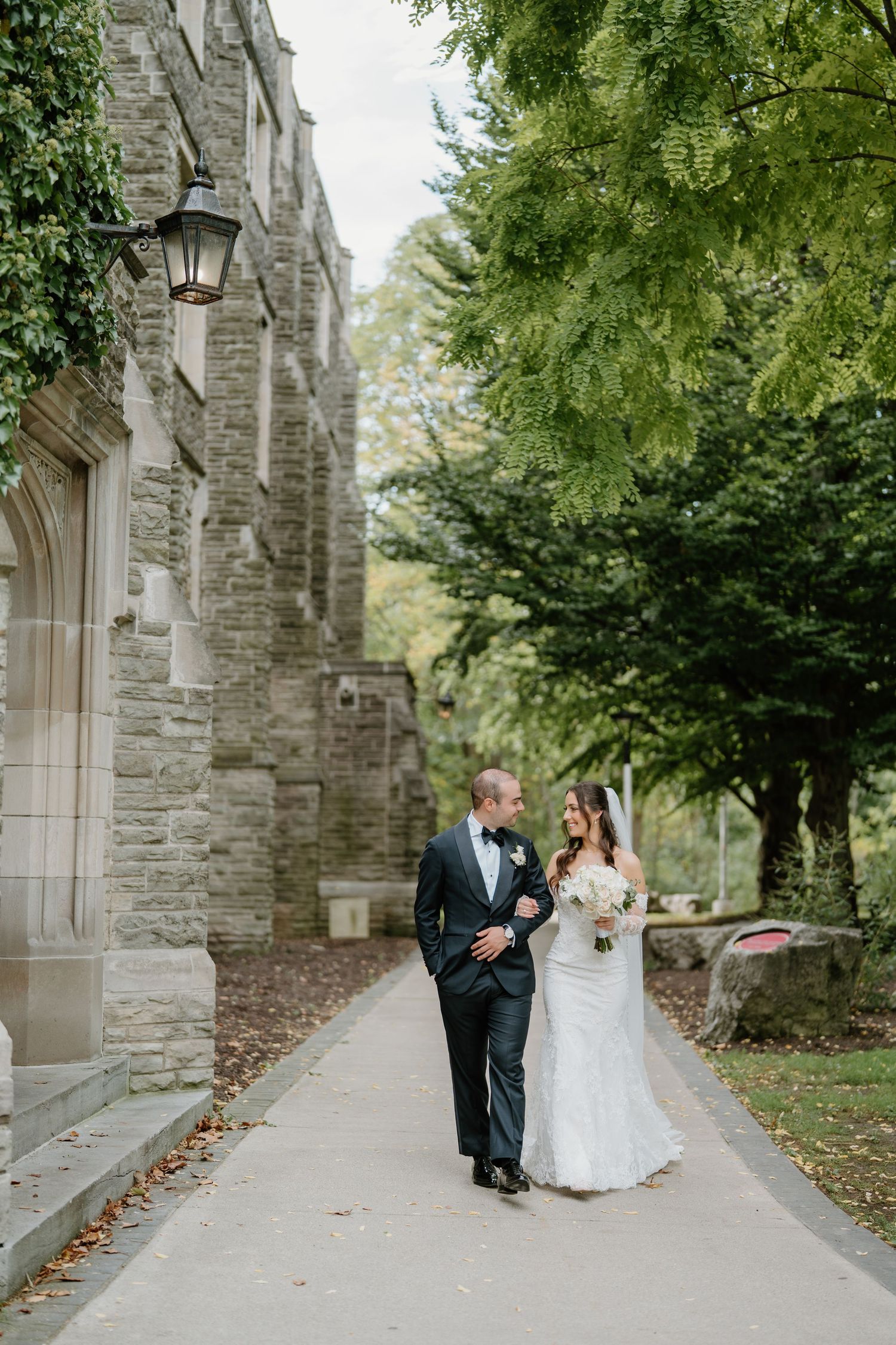 A couple walks along a stone church pathway surrounded by green trees and gothic architecture on their wedding day.