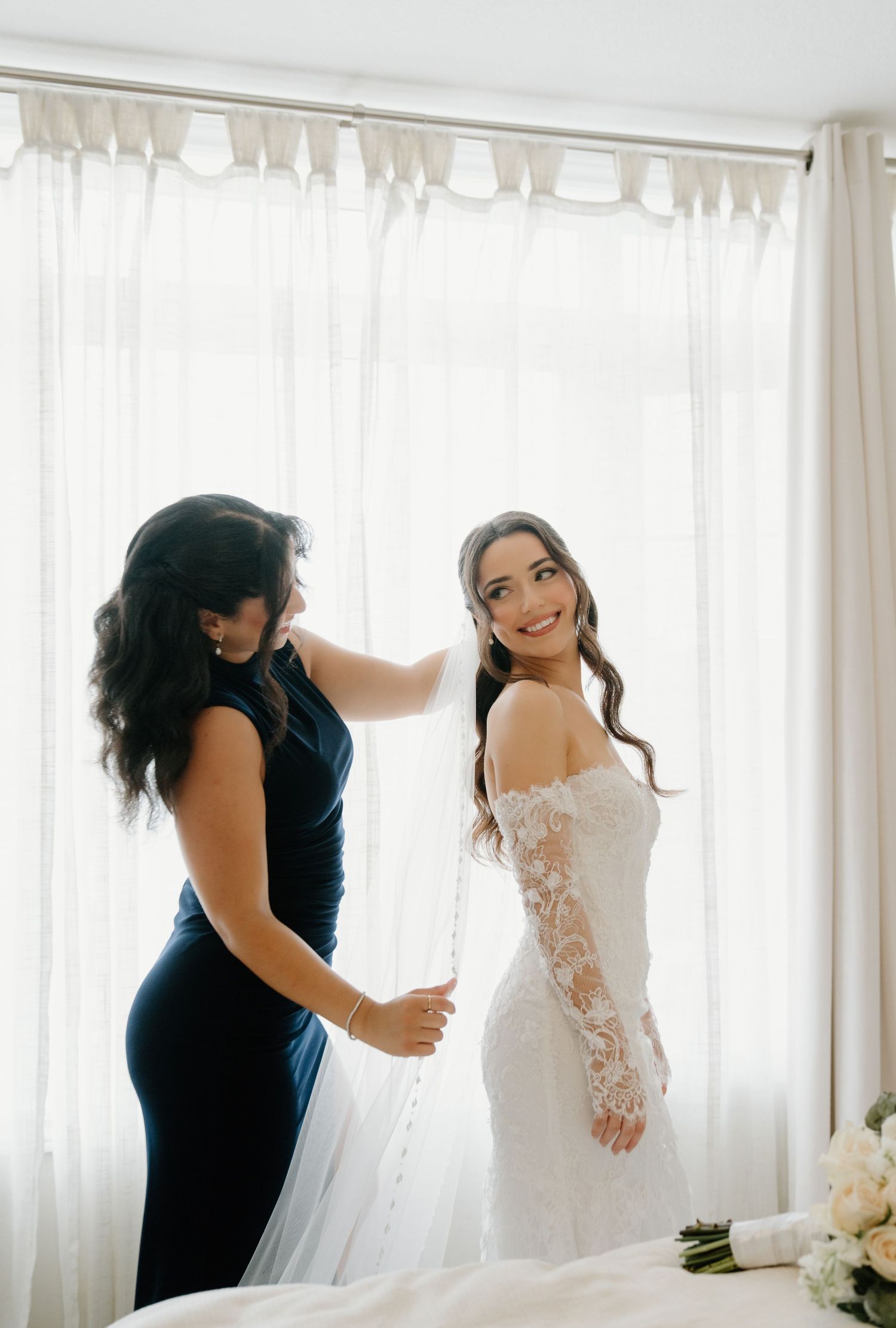 A bride in a lace long-sleeve wedding dress gets help with final adjustments near a bright window.