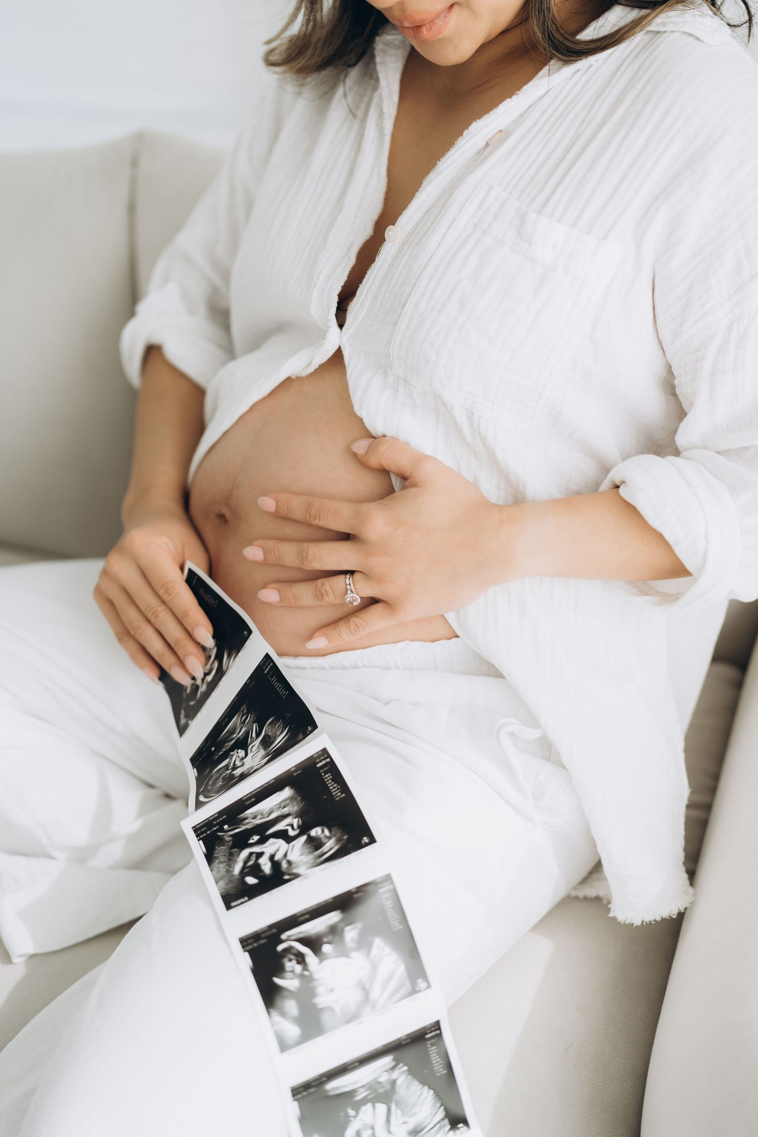 A series of ultrasound photos being held against a pregnant person's belly wearing white clothing.