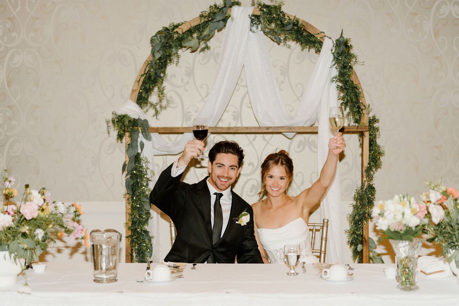 A couple celebrates under a floral arch at their wedding reception head table decorated with greenery.