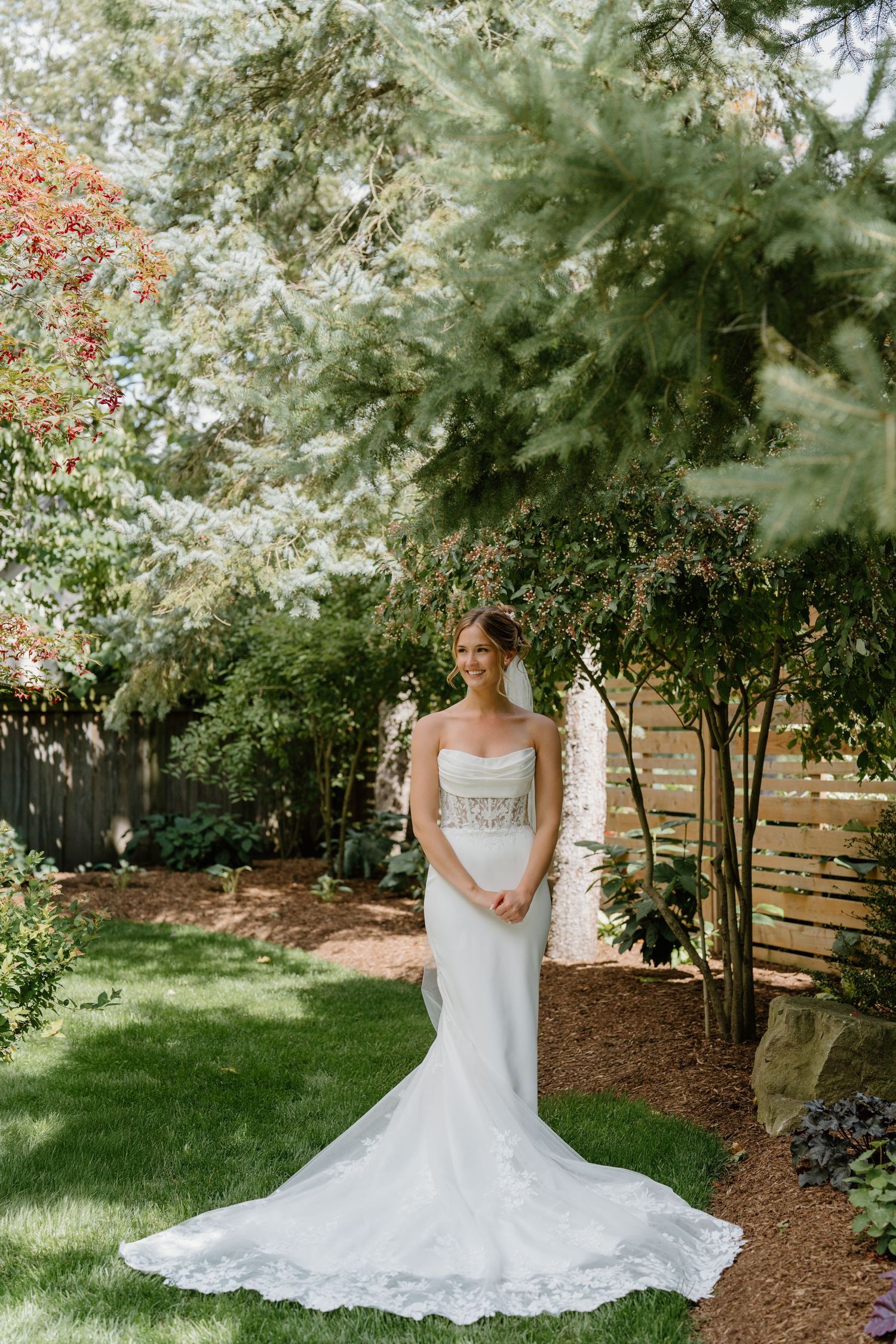 Elegant wedding dress with lace train photographed in a lush garden setting with blooming flowers and evergreen trees.