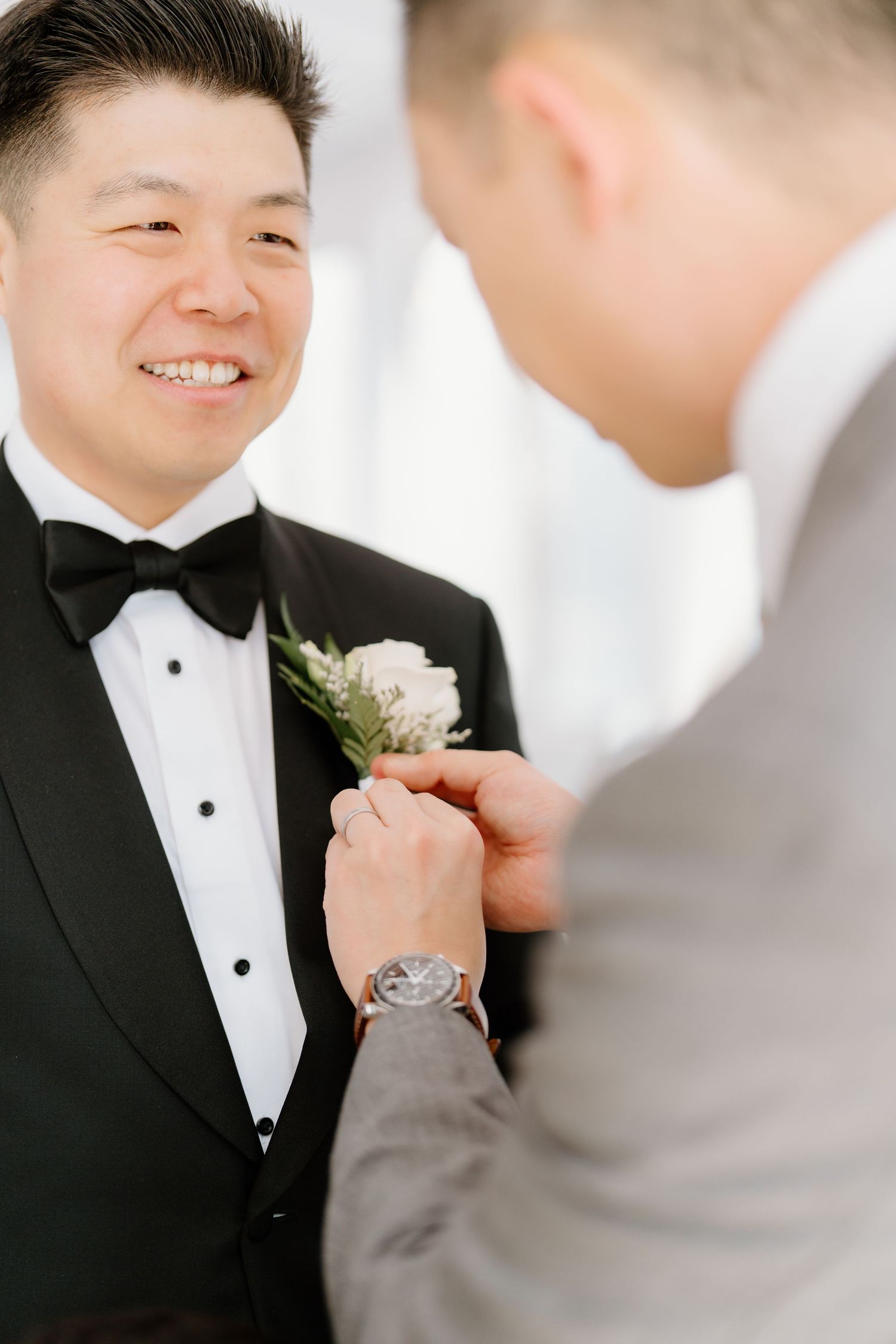 Groomsman pins white boutonniere flower onto smiling groom's black tuxedo lapel during wedding preparation.