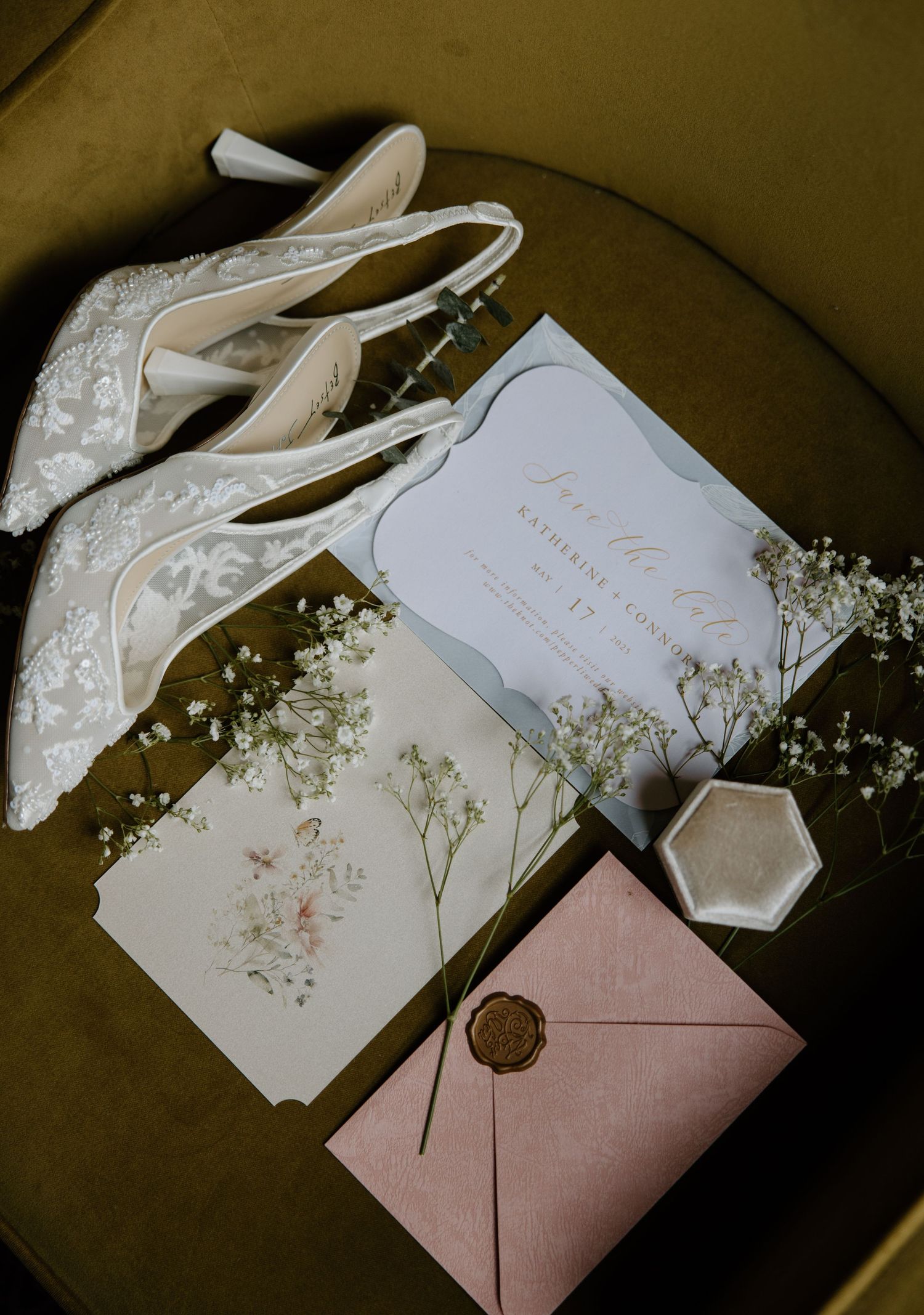 Elegant white lace wedding heels arranged with baby's breath flowers, invitation cards and a blush pink wax-sealed envelope.
