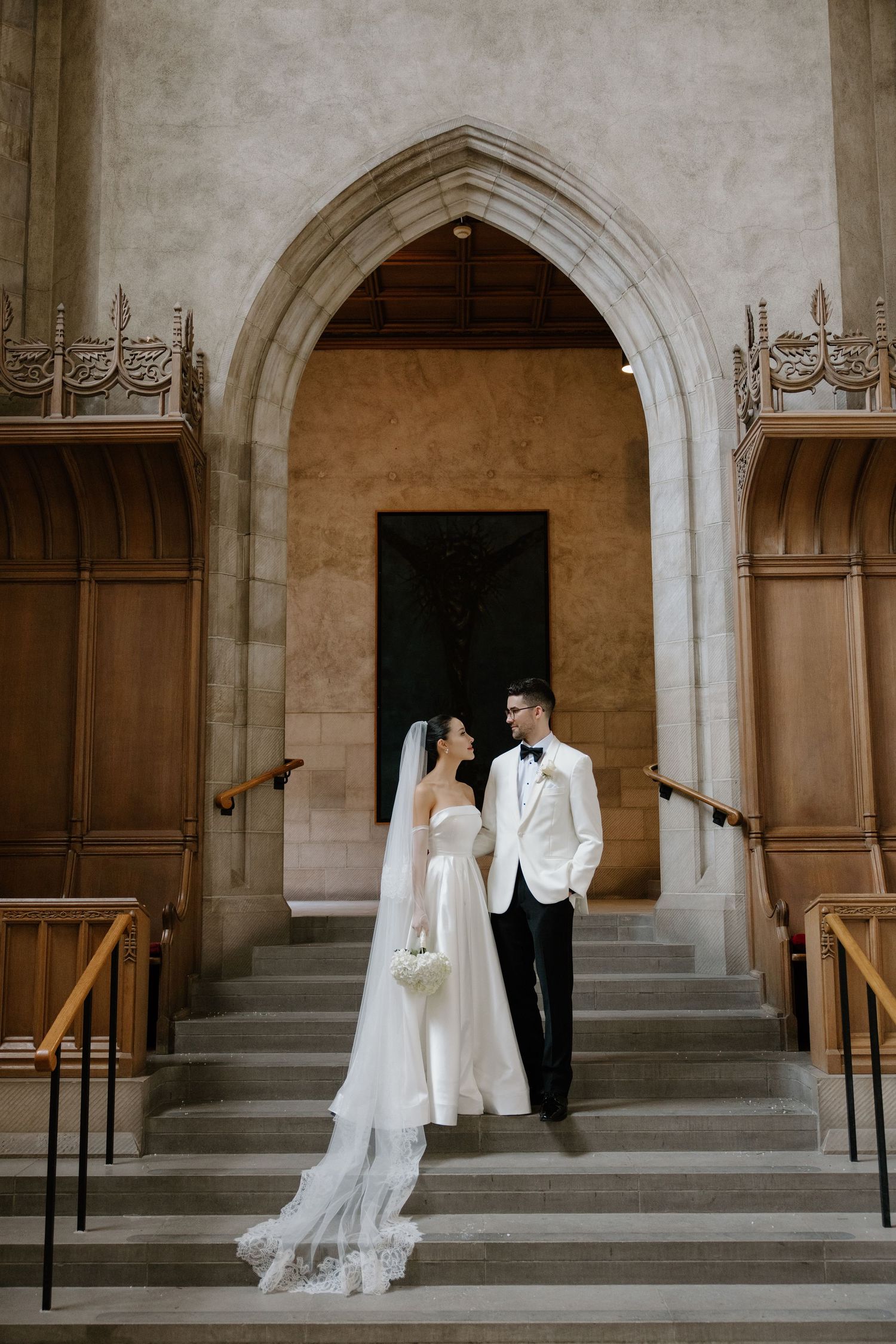 A couple stands elegantly on stone steps beneath a grand Gothic archway in a church during their wedding ceremony.