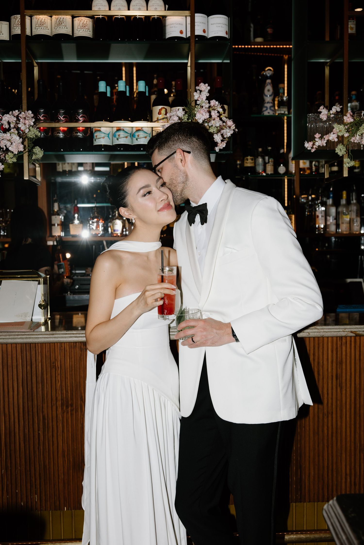 A couple in white formal attire shares an intimate moment while holding cocktails at an elegant bar.