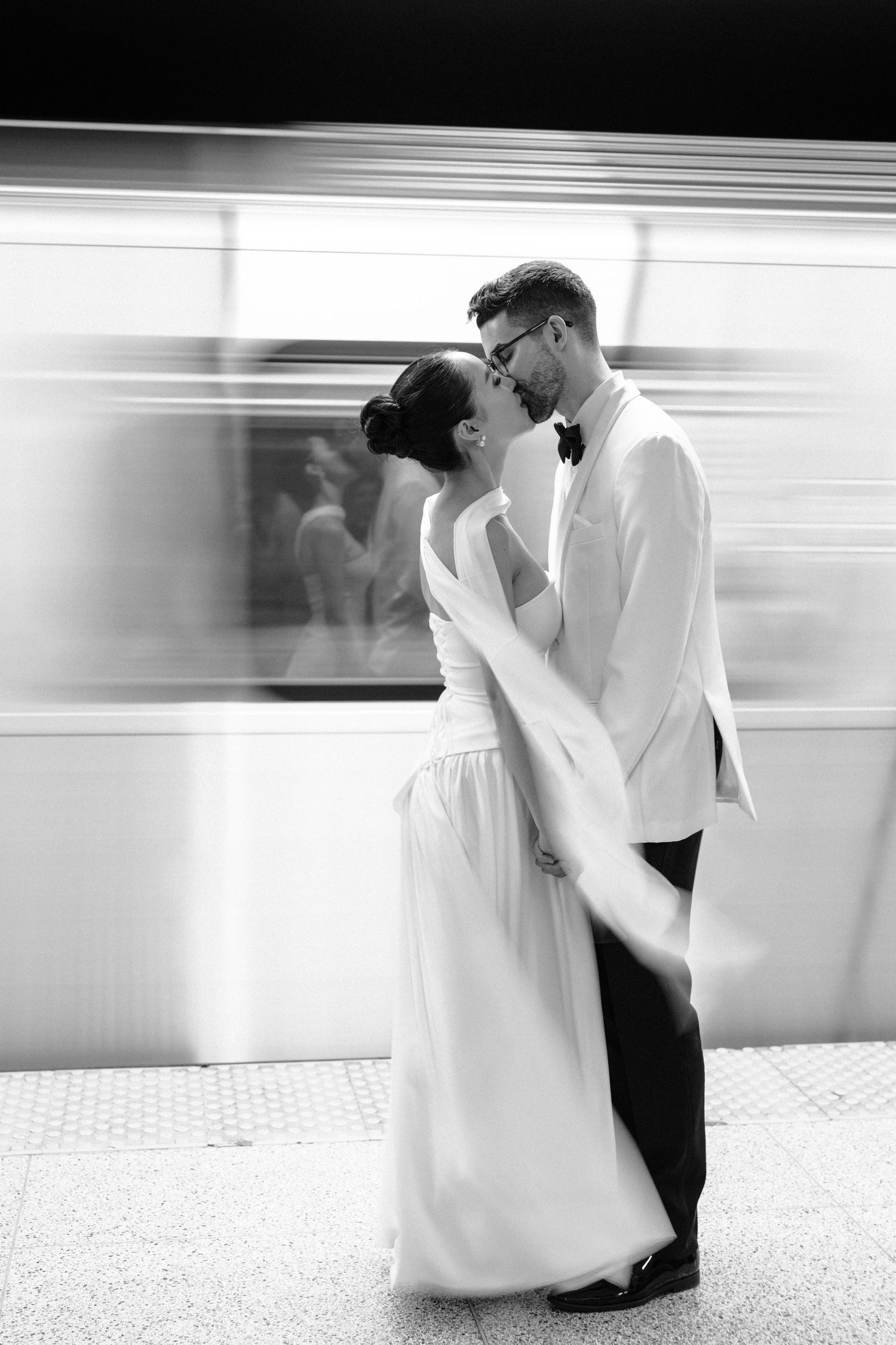 Black and white wedding photo showing a romantic couple embracing on a train platform with motion blur in background.