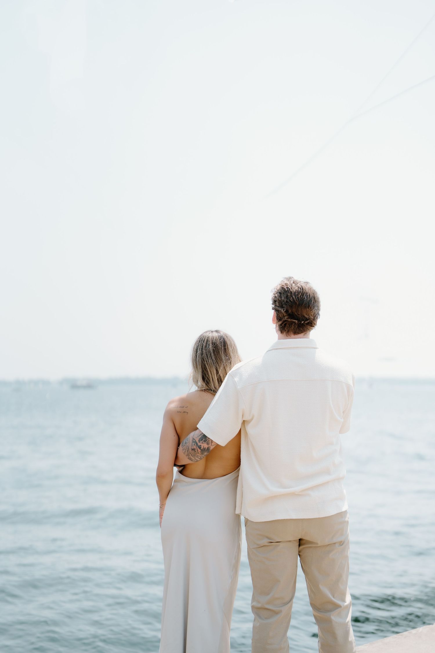 A couple stands together at the ocean's edge, embracing while looking out at the serene blue water.