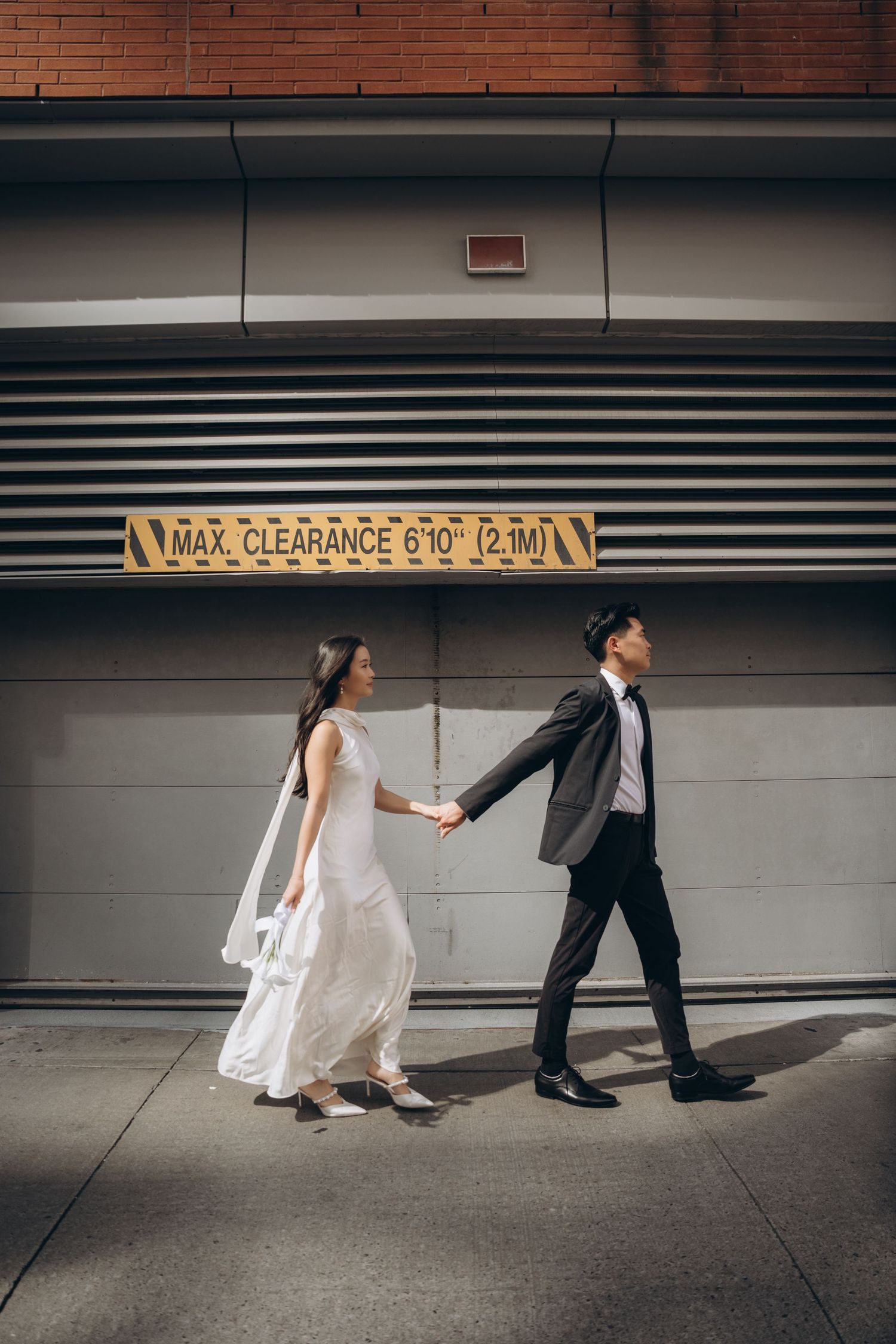 A couple in wedding attire holds hands and dances playfully along a modern concrete wall in an urban setting.