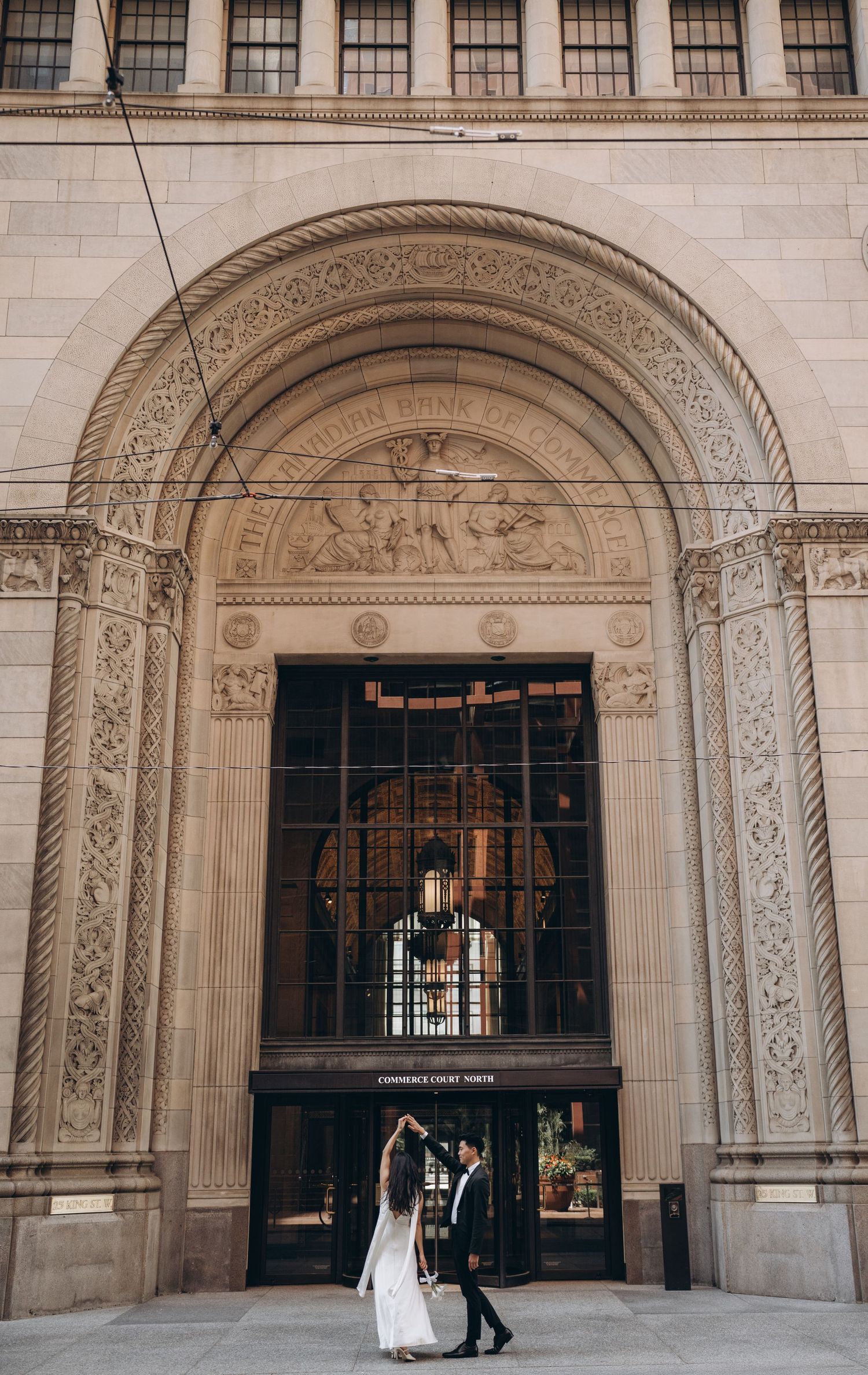 A couple in wedding attire walks through an ornate stone archway in a historic building with architectural details.