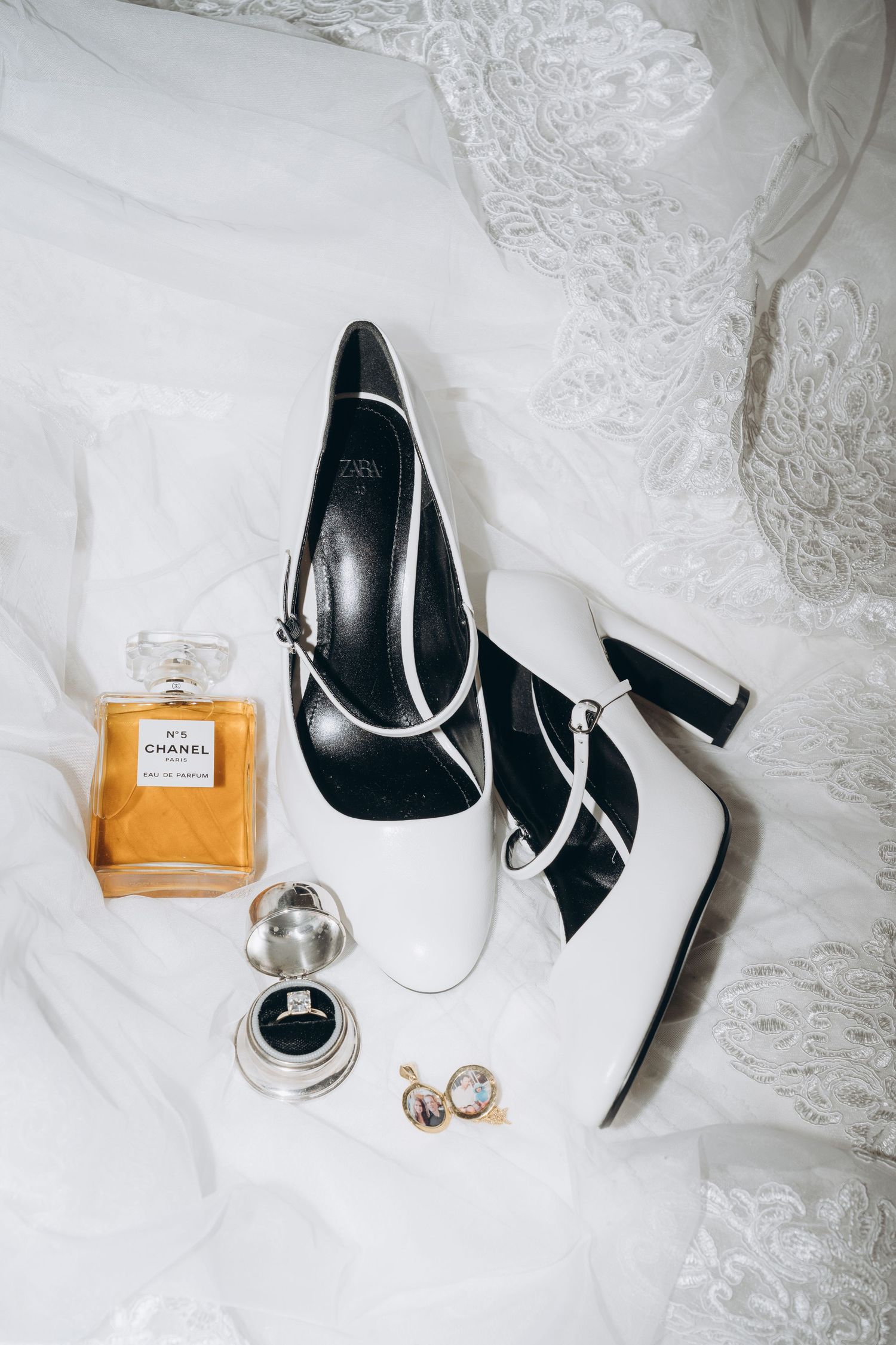 Wedding accessories with perfume bottle, black and white shoes, and jewelry on white lace background.