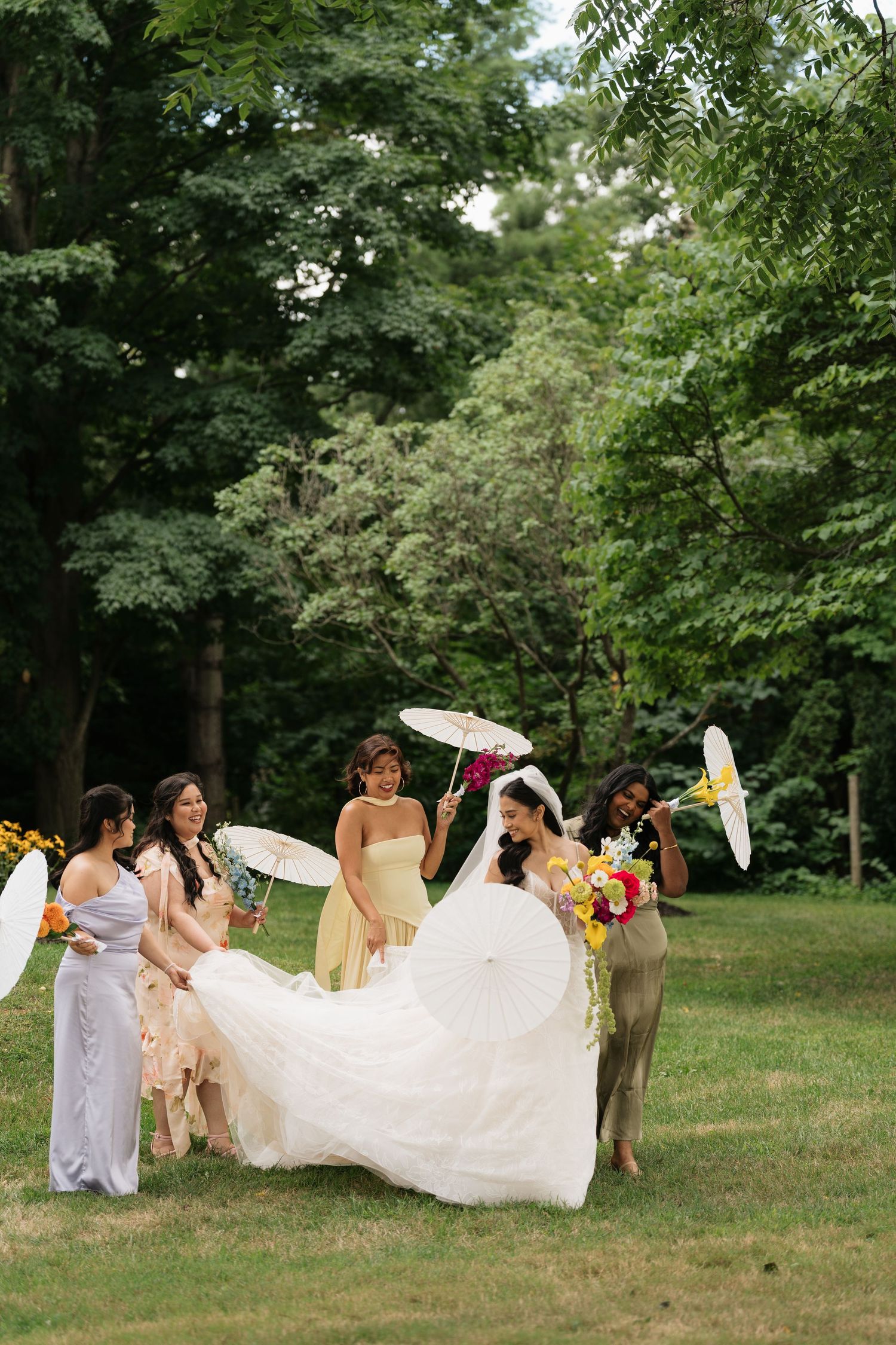A bridal party with parasols gathers on a lush green lawn surrounded by trees on a sunny summer day.