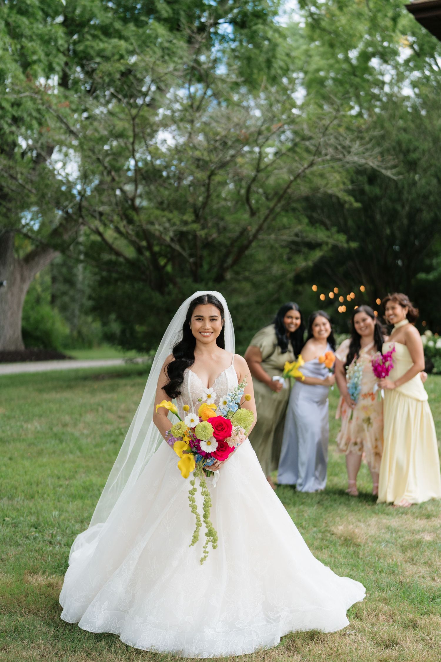 A bride in a white ball gown poses with her colorful bouquet while bridesmaids gather on a grassy lawn.