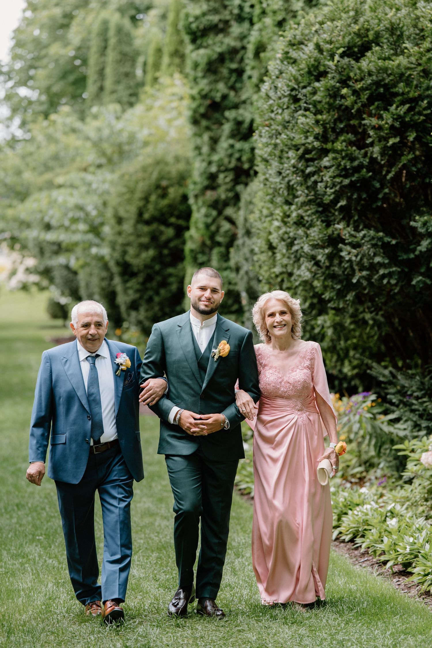 Three people walking together on a green lawn path bordered by garden shrubs and flowers in formal attire.