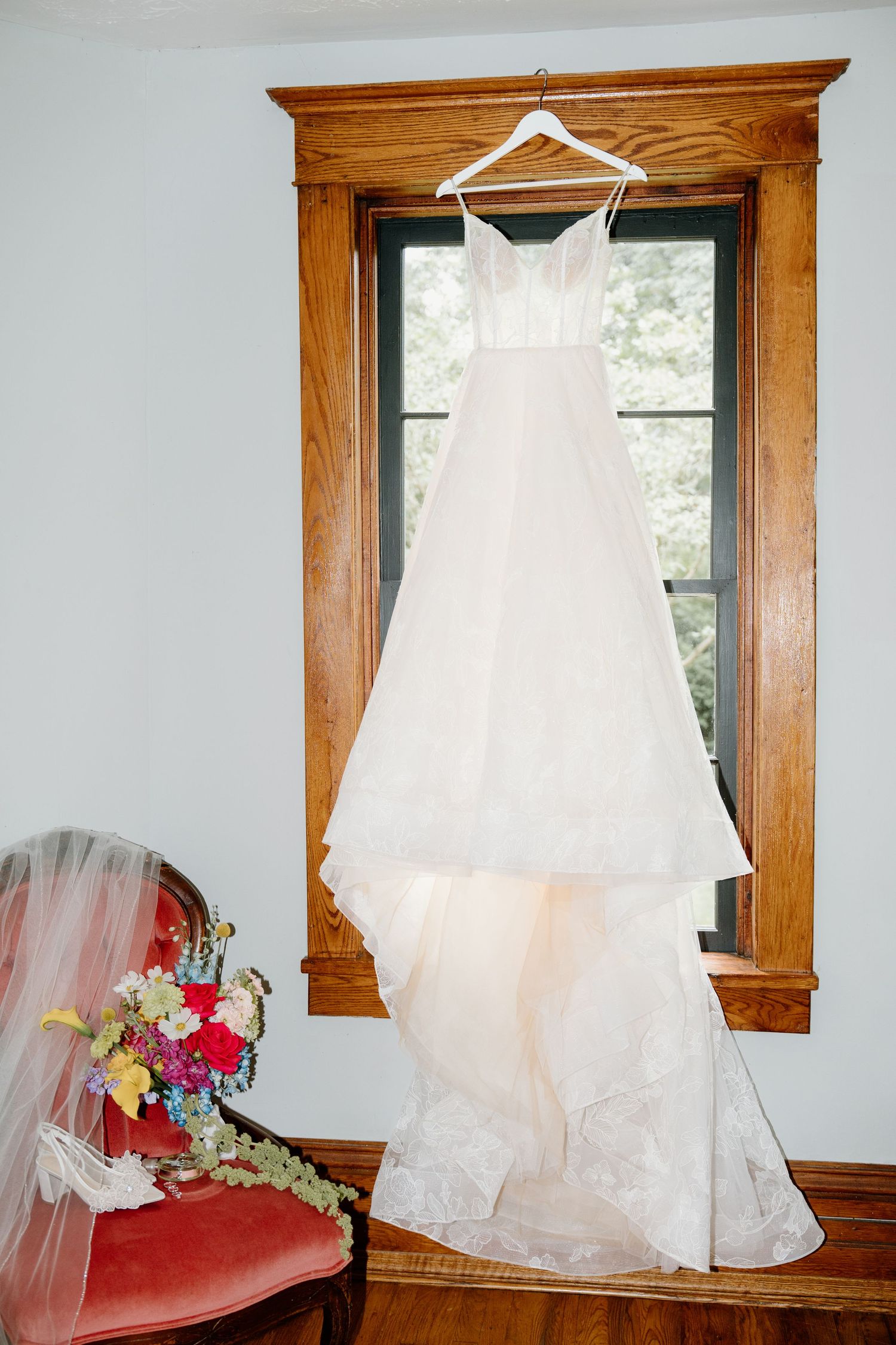 A white wedding dress hangs elegantly by a window with a wooden frame and red floral bouquet arrangement.