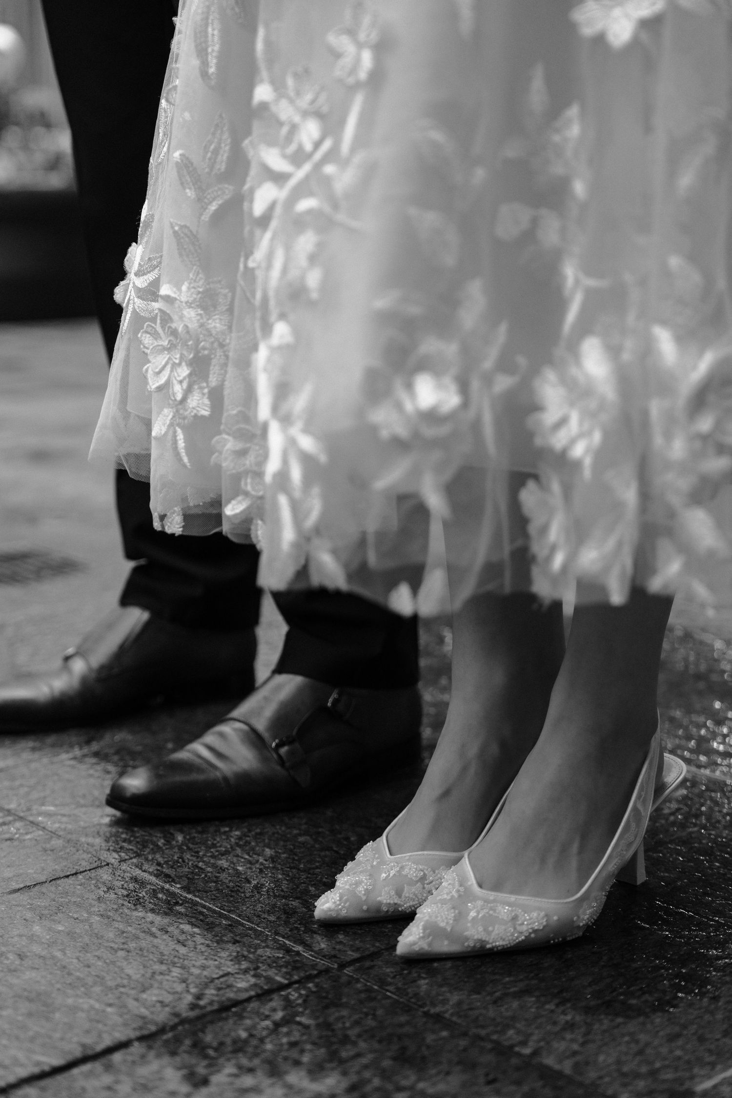 Black and white wedding photo showing bride's white lace dress and flats next to groom's black dress shoes on stone floor.